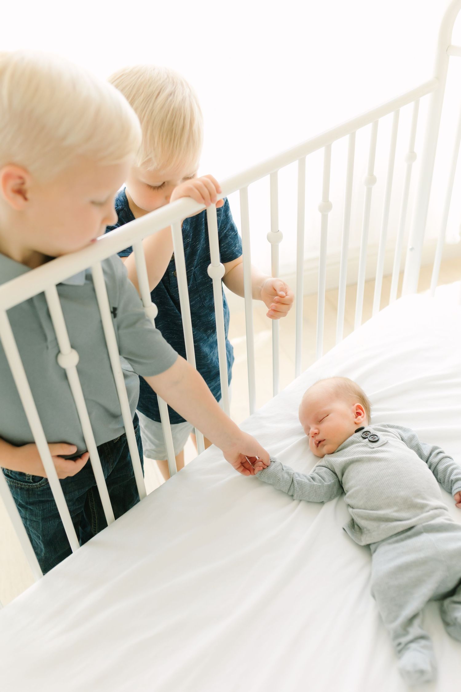Two blonde toddlers gently touch their sleeping newborn sibling in a white crib, sharing a tender family moment.