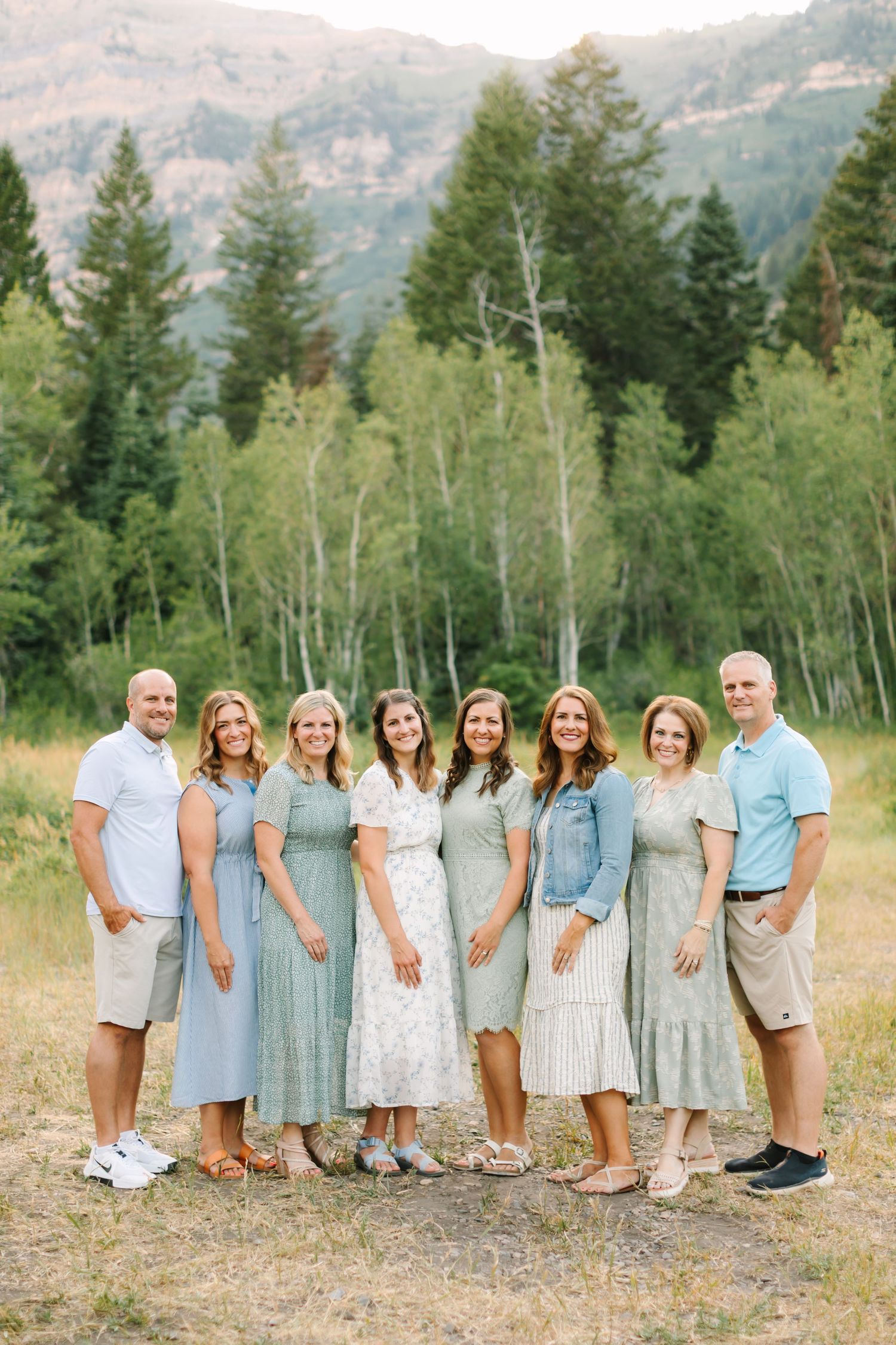 Large family group smiling outdoors in coordinated sage green and white outfits with lush green trees behind in Provo, Utah.