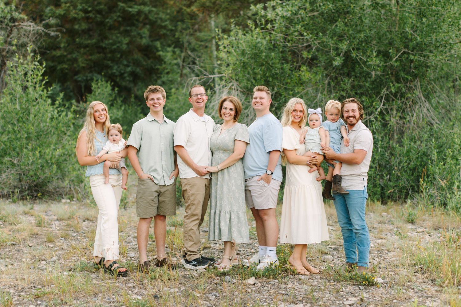Large family group smiling outdoors in coordinated sage green and white outfits with lush green trees behind in Provo, Utah.