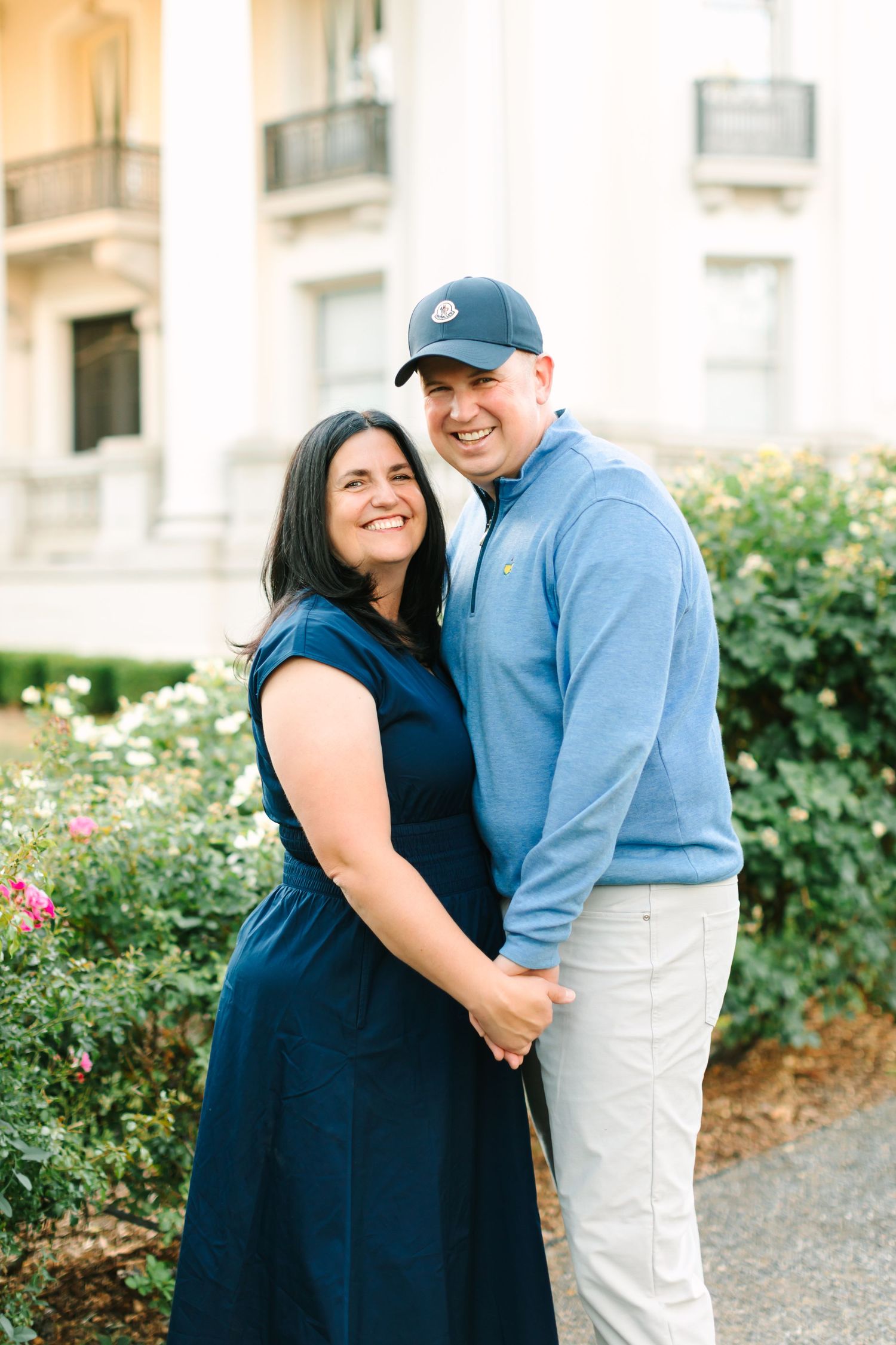 Smiling couple holding hands outdoors near flowering bushes, woman in navy dress, man in blue pullover and cap.
