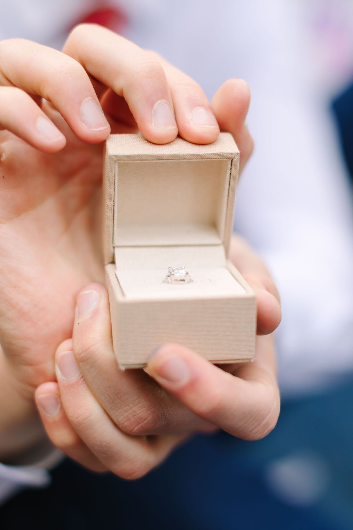 Hands holding open a beige ring box with a diamond solitaire engagement ring inside.