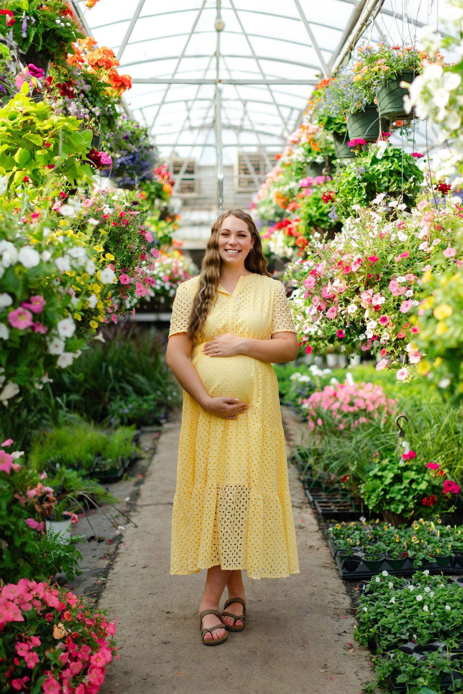 A person in a yellow polka dot dress poses joyfully among colorful flowers in a greenhouse setting.