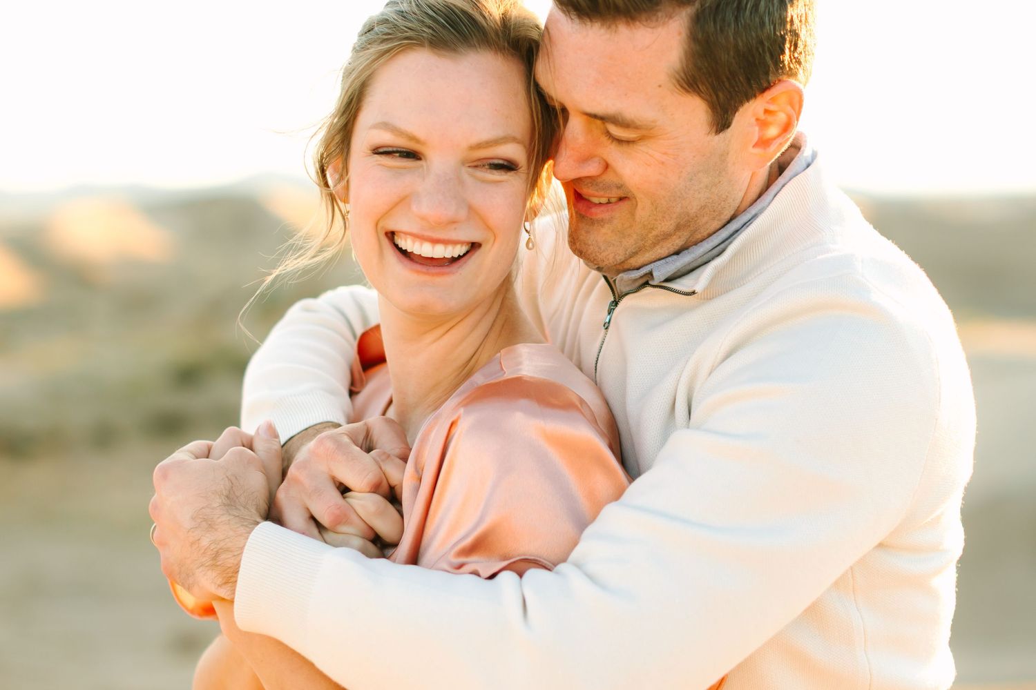 A romantic couple embraces and laughs together during a golden hour photoshoot in a natural outdoor setting.