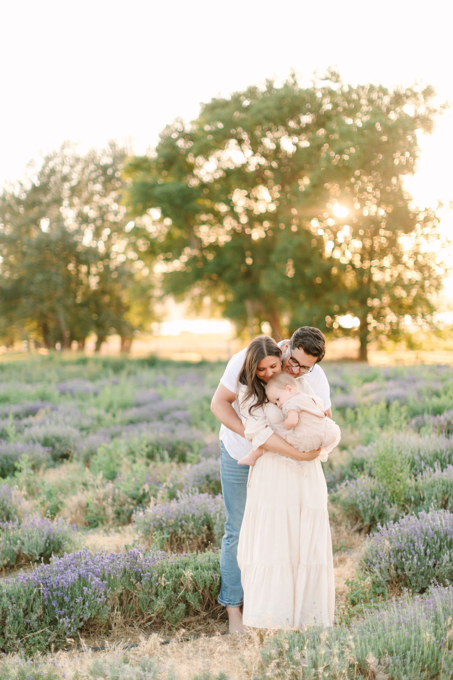 A couple embraces in a beautiful lavender field at sunset with golden light filtering through trees in the background.