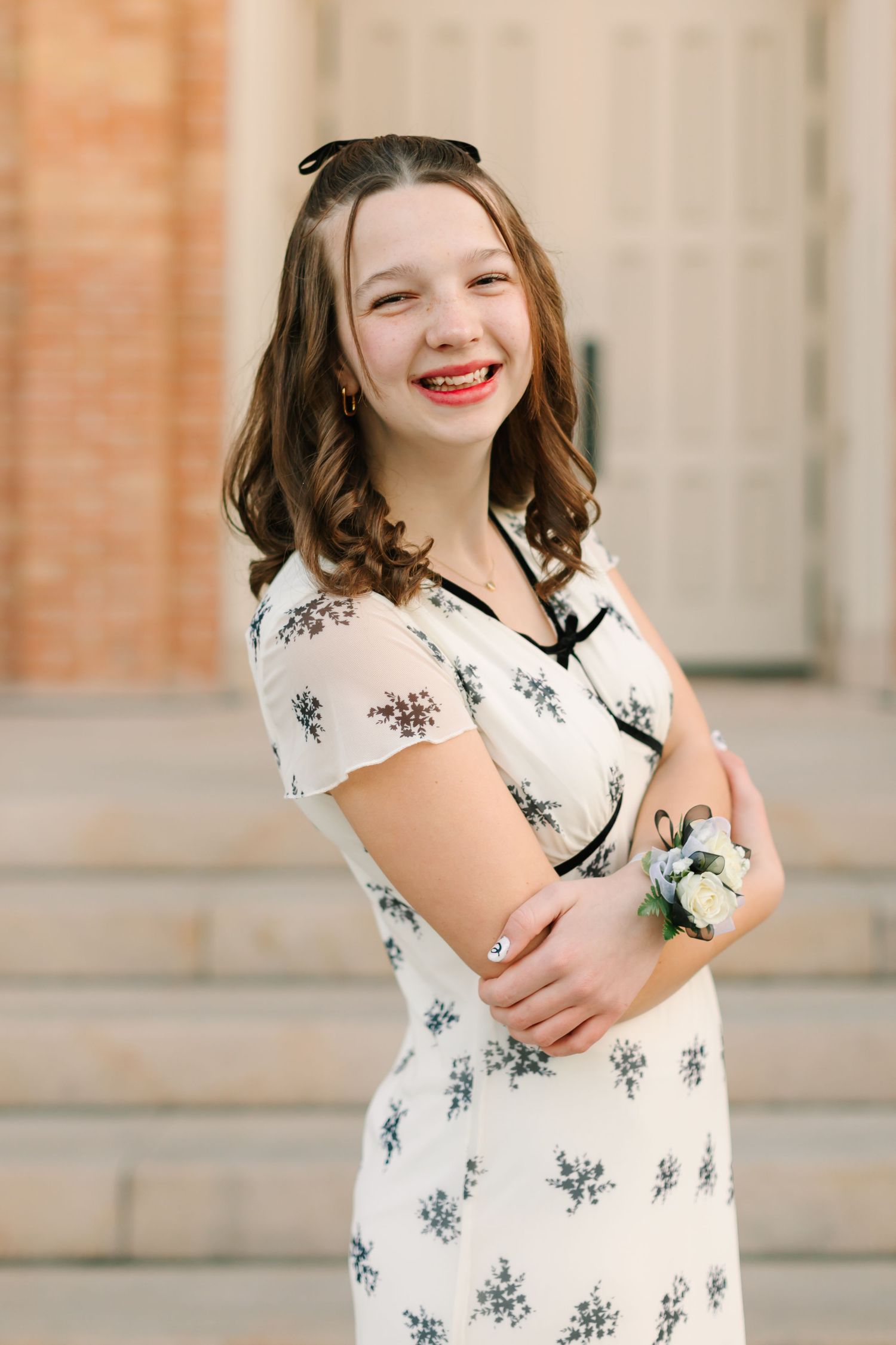 A person in a floral white dress smiles while wearing sunglasses on their head and holding a corsage.