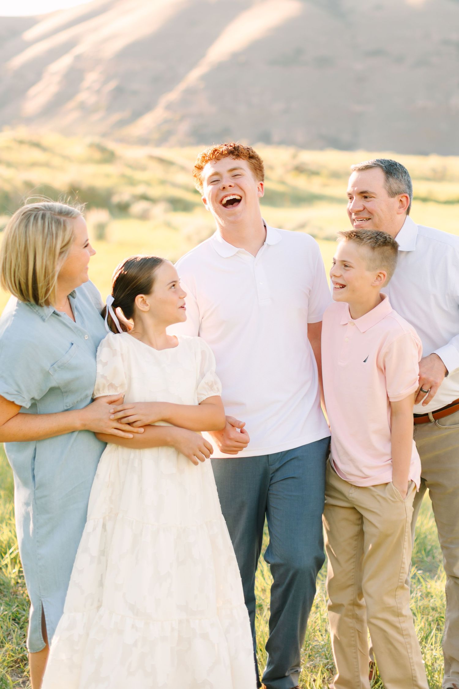 A joyful family of six laughing together outdoors in a sunny field with rolling hills in the background.