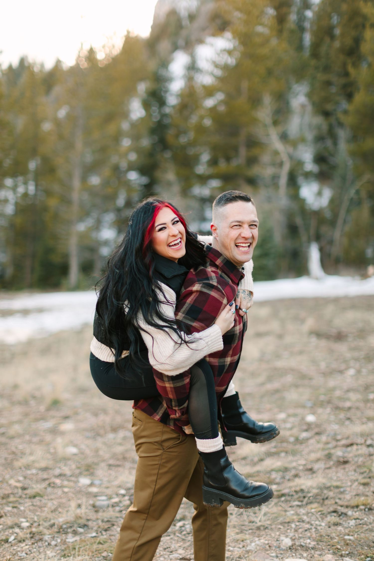 A couple shares a joyful piggyback ride moment in a winter forest setting wearing casual outdoor attire.