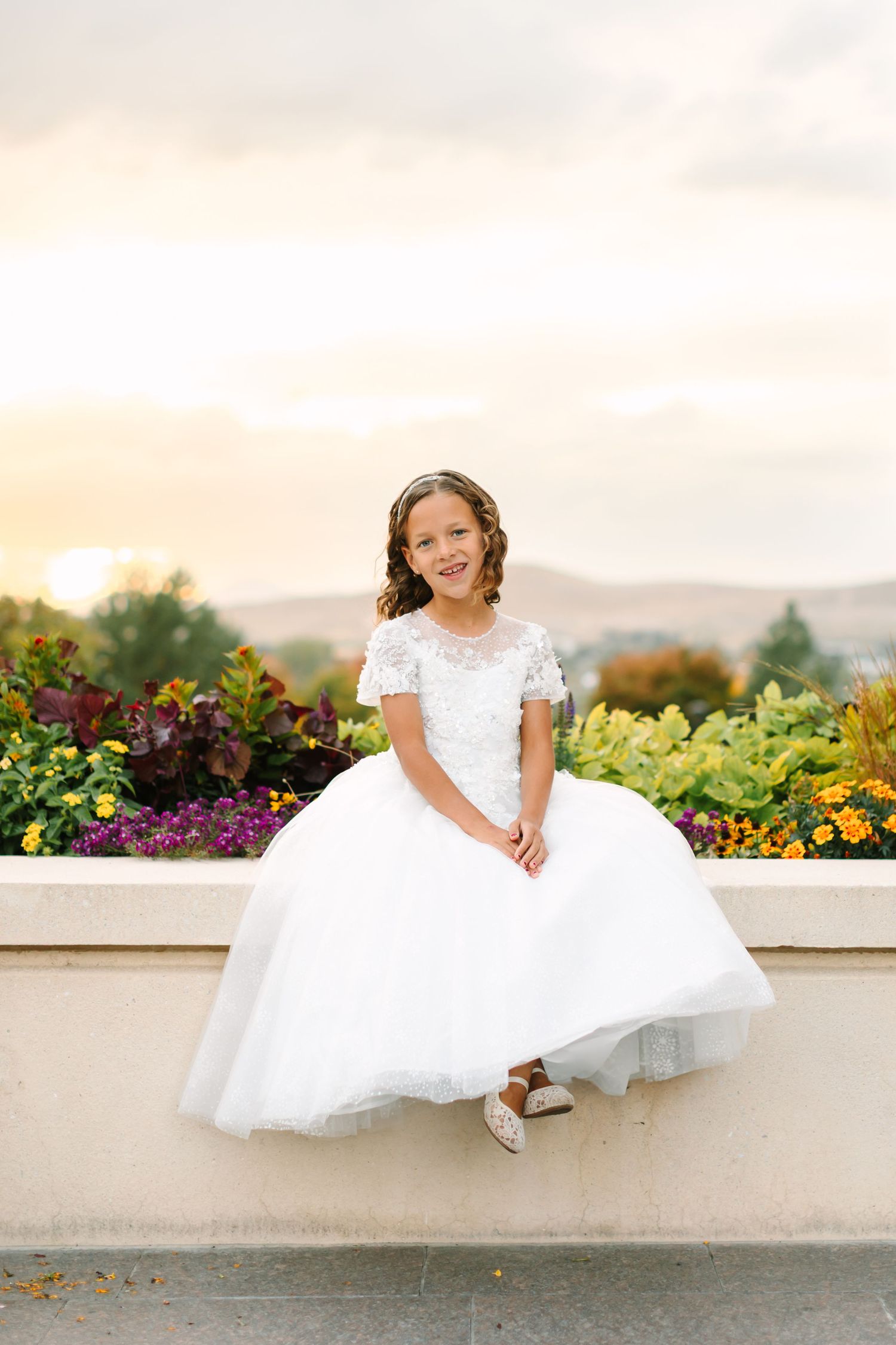 A child in a white dress sits on a ledge surrounded by colorful flowers during a sunset photoshoot.