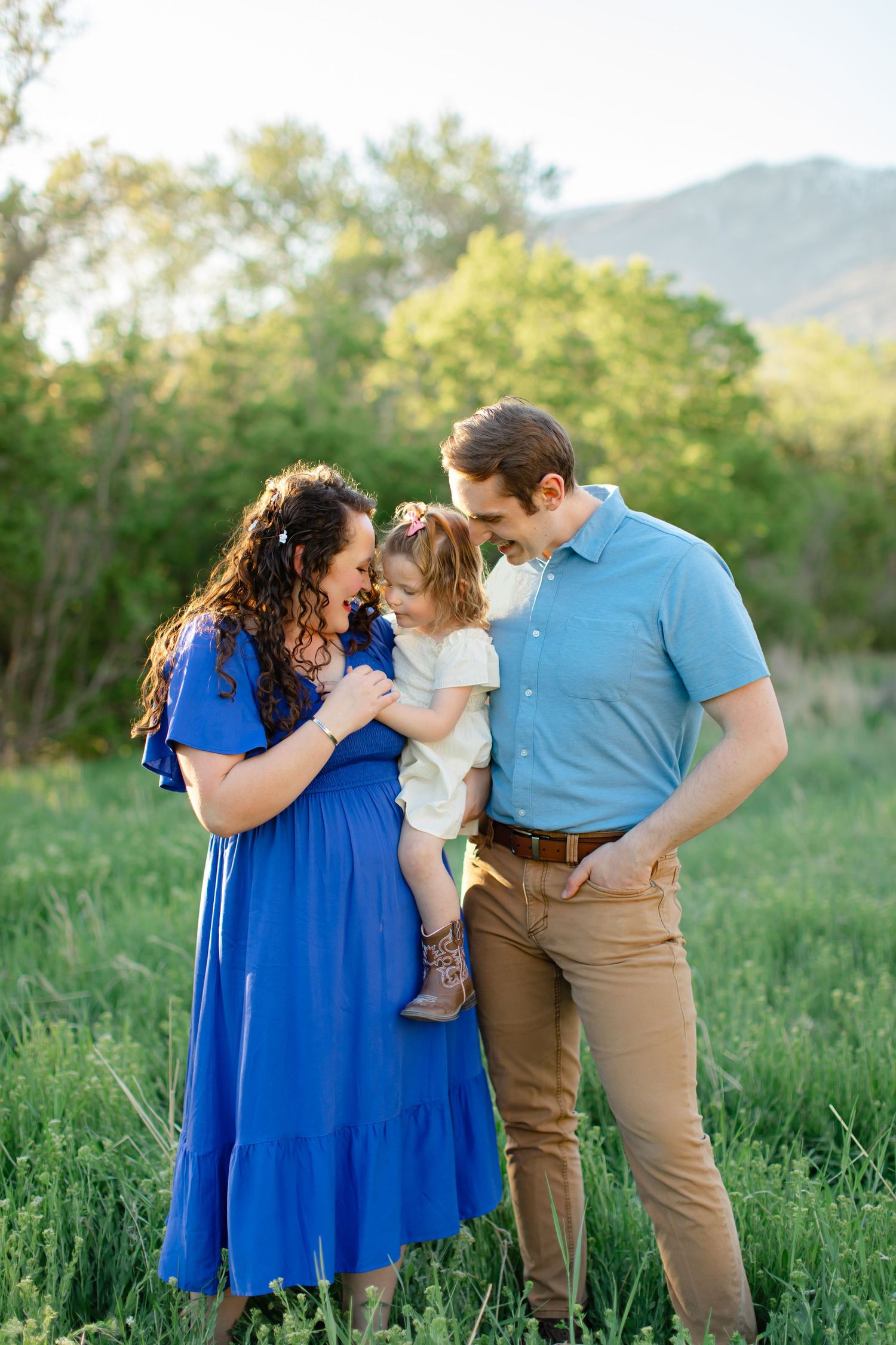 Family in coordinating blue and brown outfits share a tender moment together in a sunlit meadow with mountains behind them.