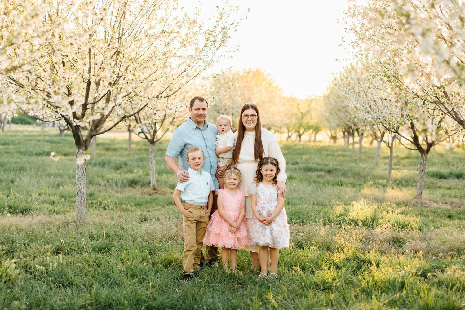 Family of six posing in a blooming orchard during golden hour, parents with four young children in spring attire in Santaquin, Utah.