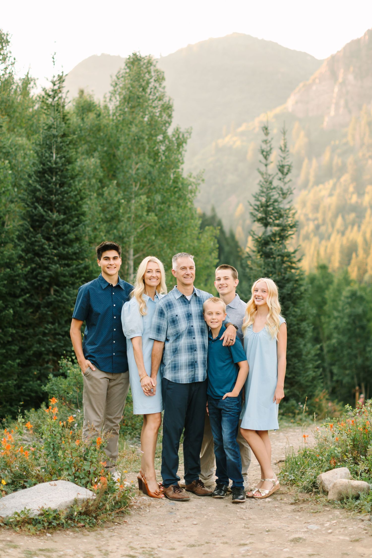 Family of six posing outdoors in mountain setting with evergreen trees, wearing coordinated blue outfits on a sunny day.
