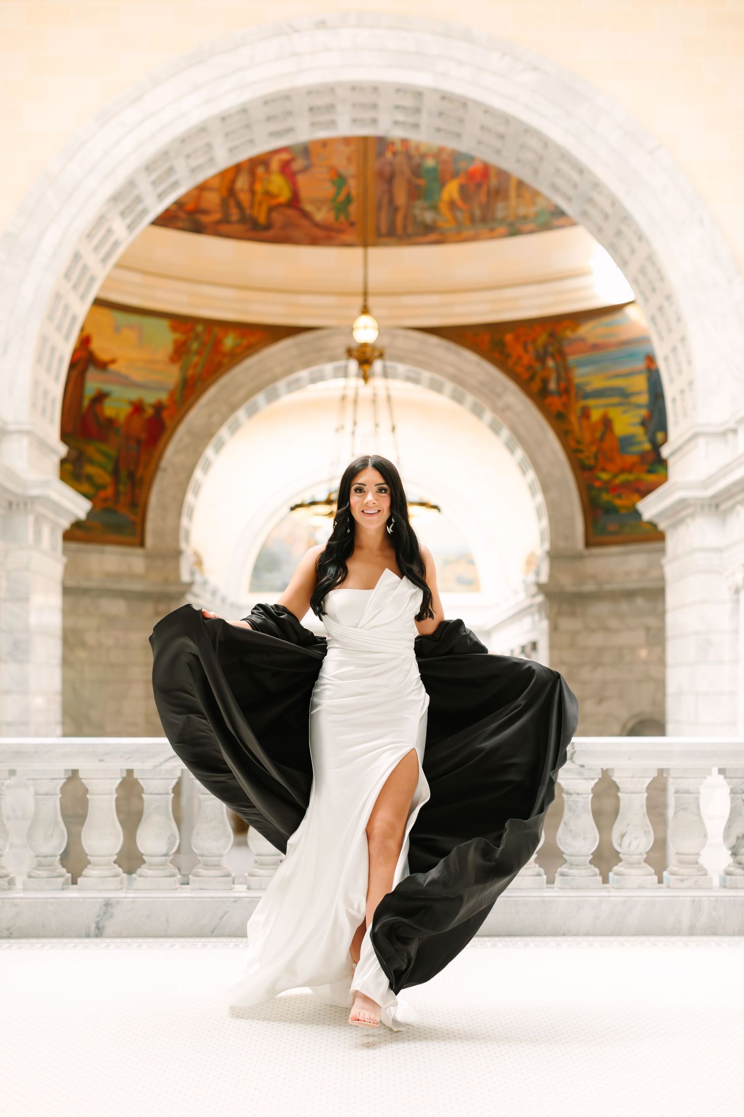 Dancer in white and black dress performs graceful poses in ornate arched hallway with painted murals and marble columns.