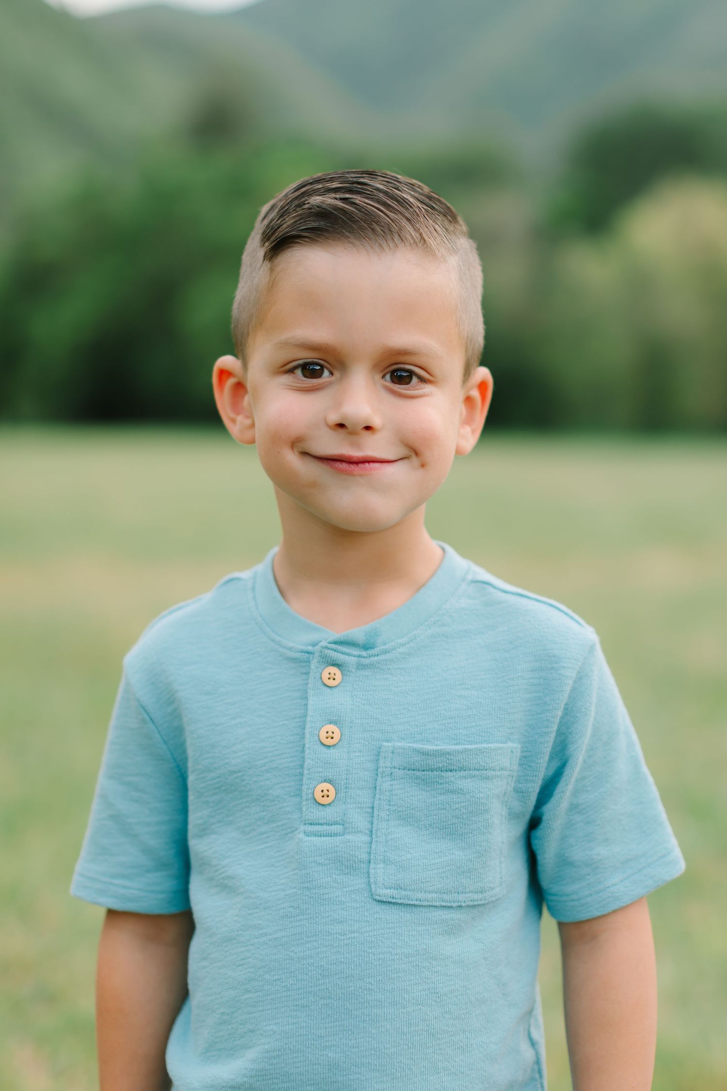 A young child wearing a light blue t-shirt with buttons stands outdoors in a grassy field on a sunny day.