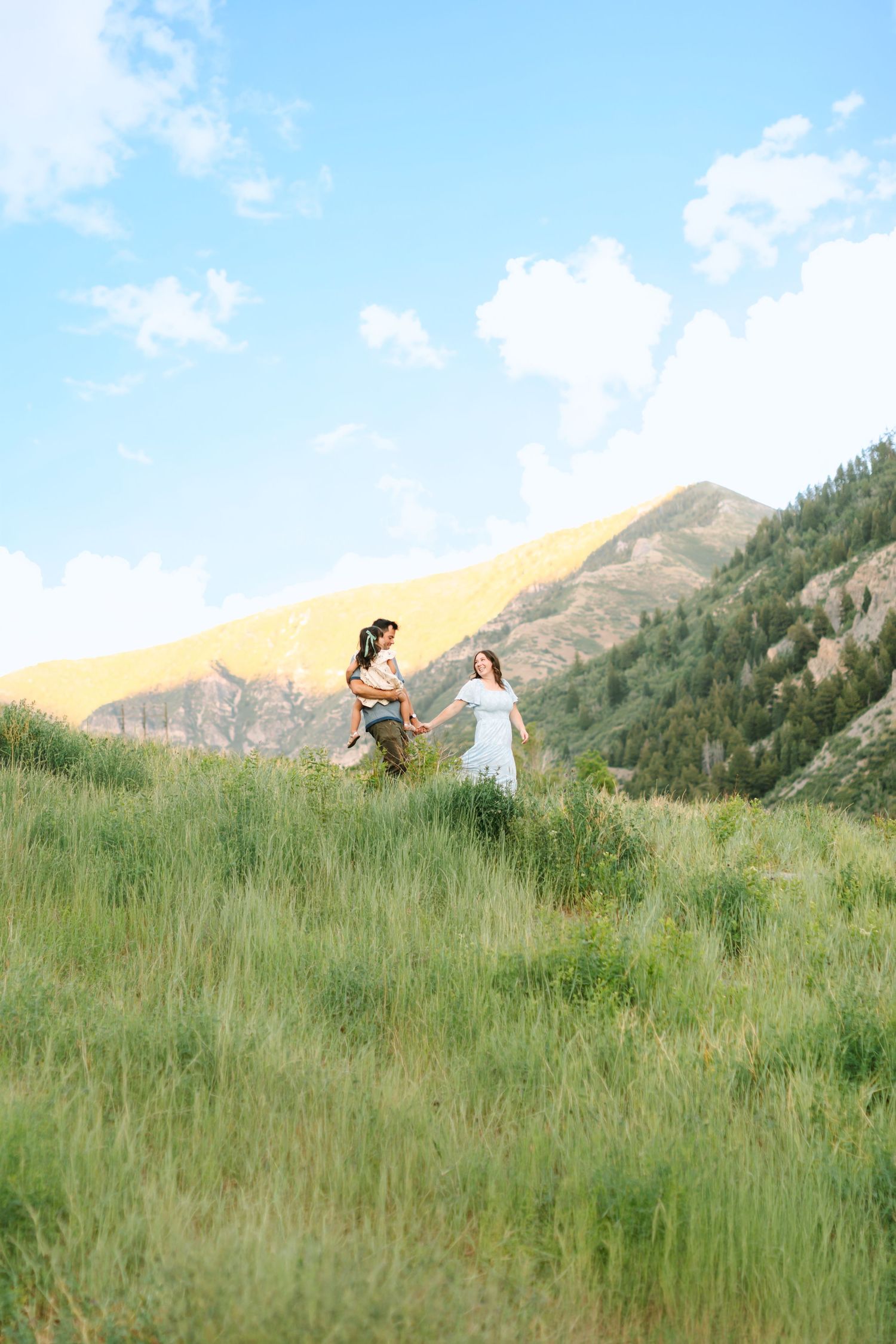 Two women in white dresses hold hands running through green mountain meadow under blue sky.