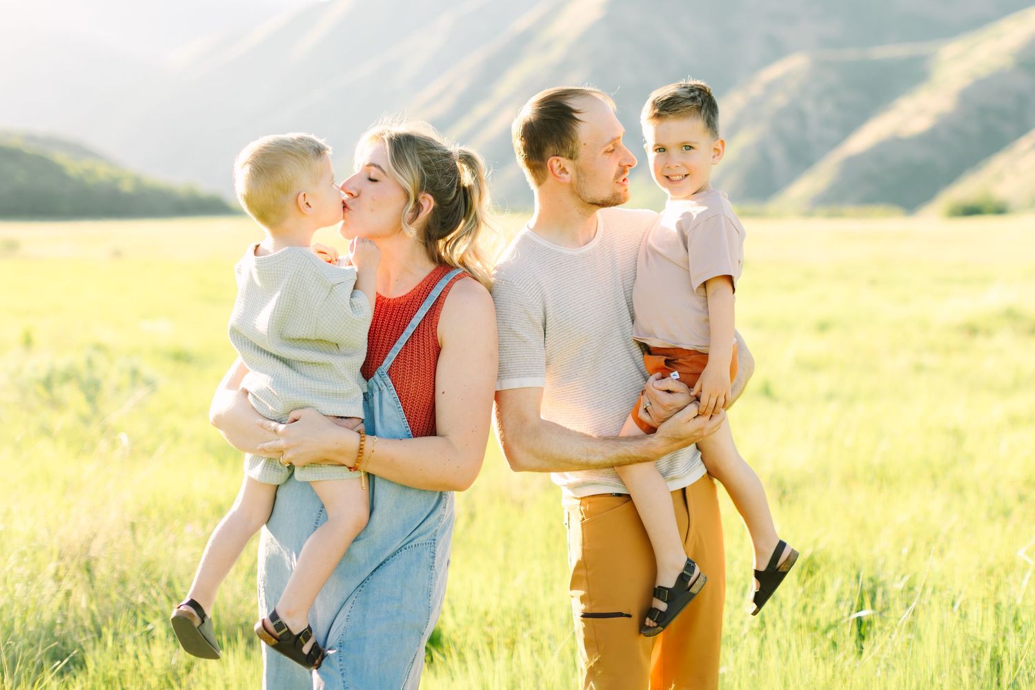 Happy family with two young boys poses in a sunny green meadow with mountain backdrop in warm golden light.