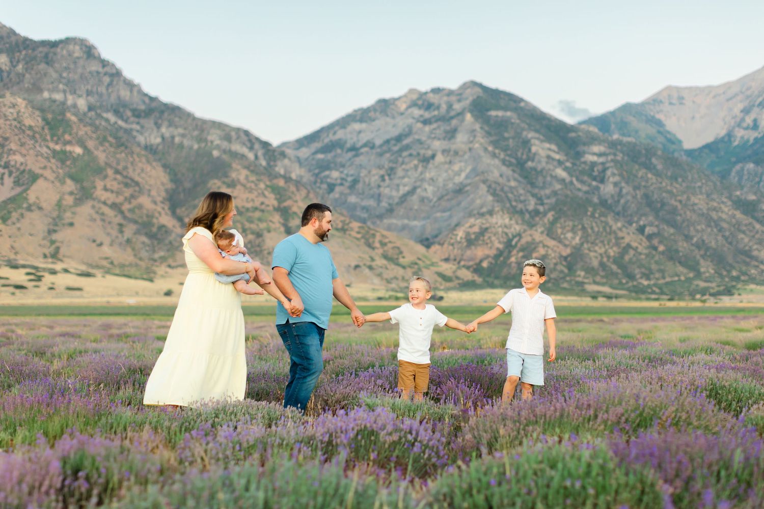 Family in white and blue outfits dancing together in a field of purple wildflowers with mountains in the background.
