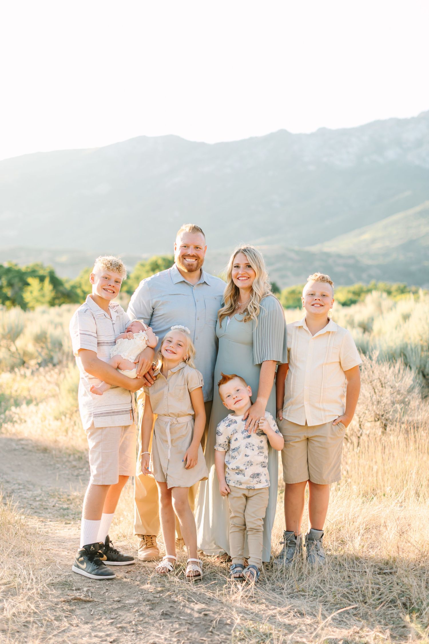 Family of six poses together on a mountain trail at sunset with golden grass and mountains in the background.