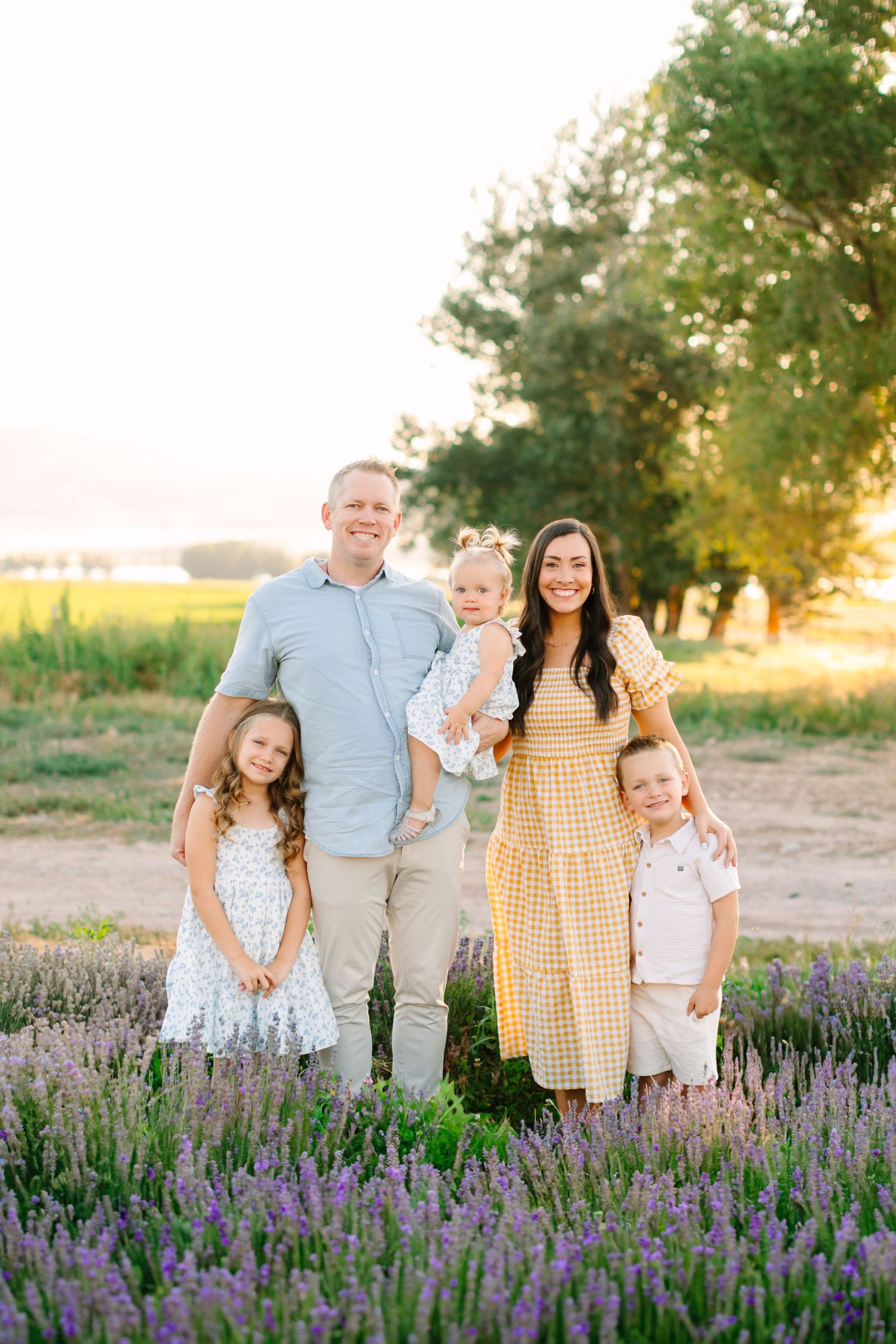 Family stands together in a beautiful lavender field at sunset with golden light filtering through the trees.