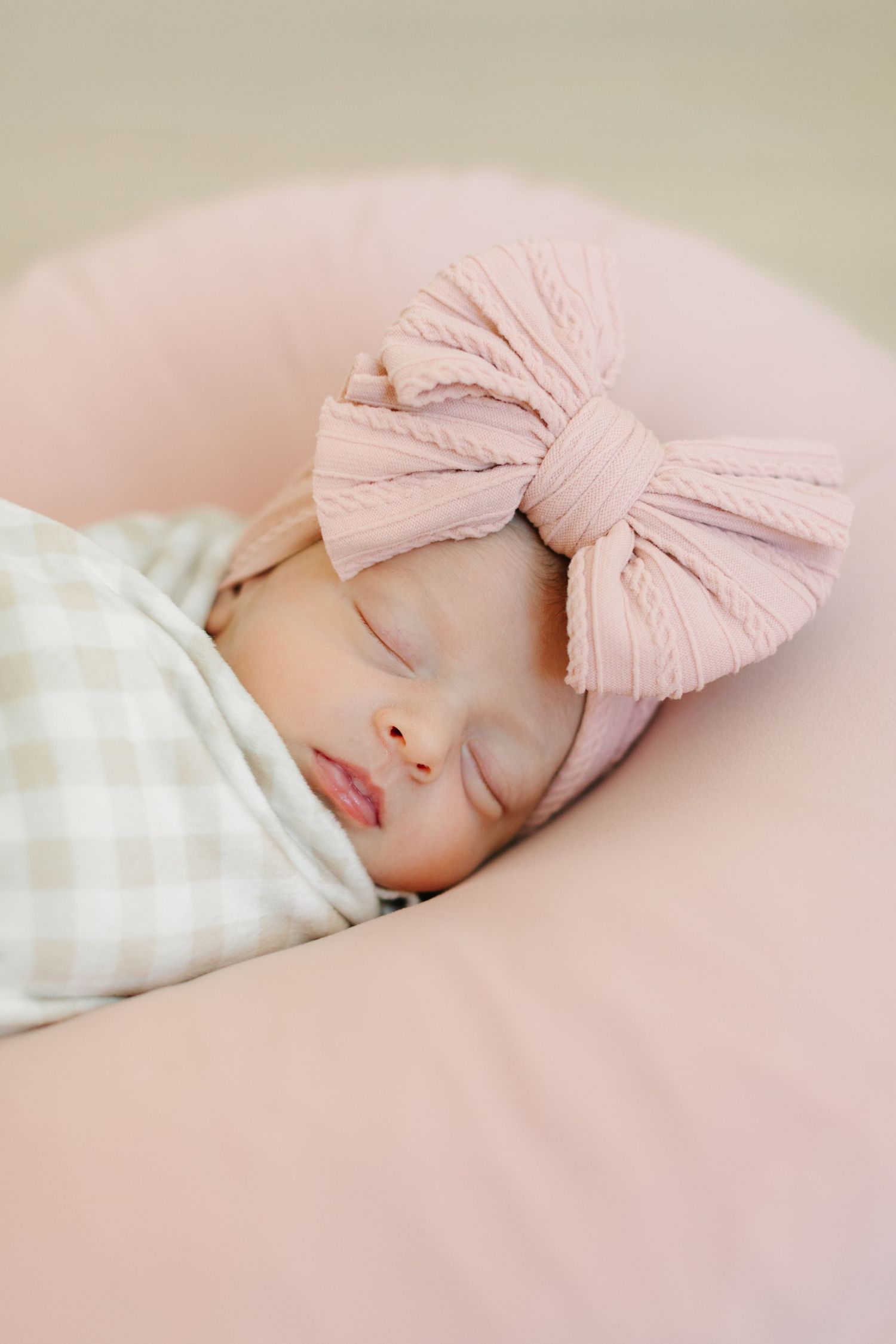 A sleeping baby wearing a soft pink bow headband rests on a blush-colored cushion.