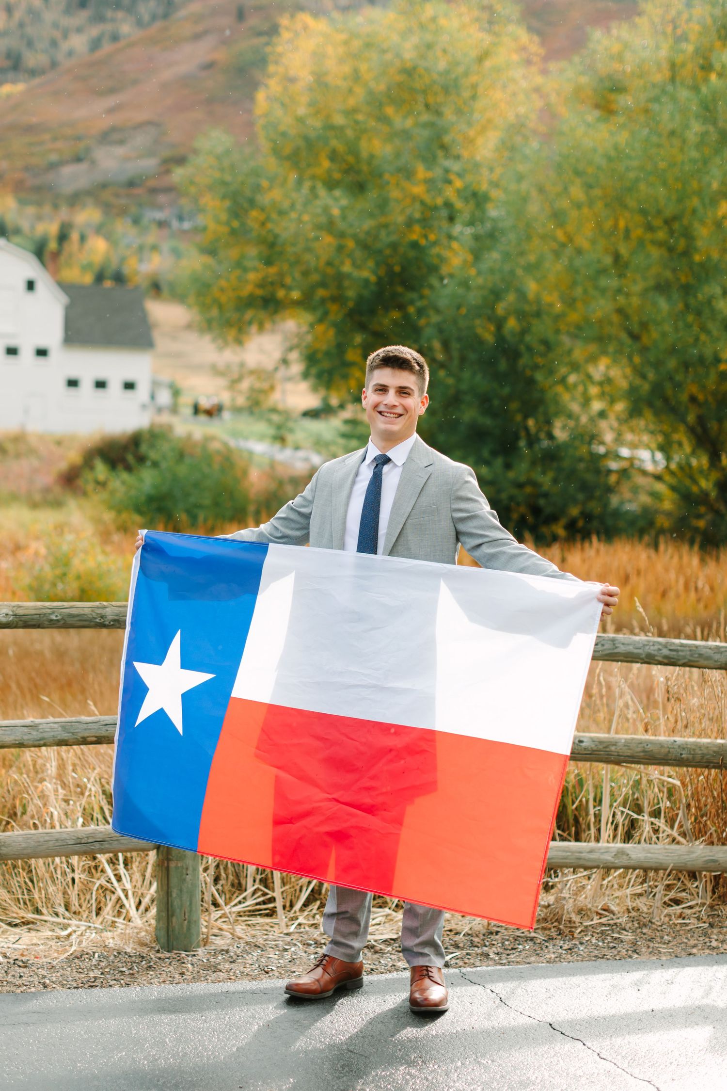 Someone in a grey suit holds a Texas state flag while standing on a road with autumn foliage and a white barn in the background.
