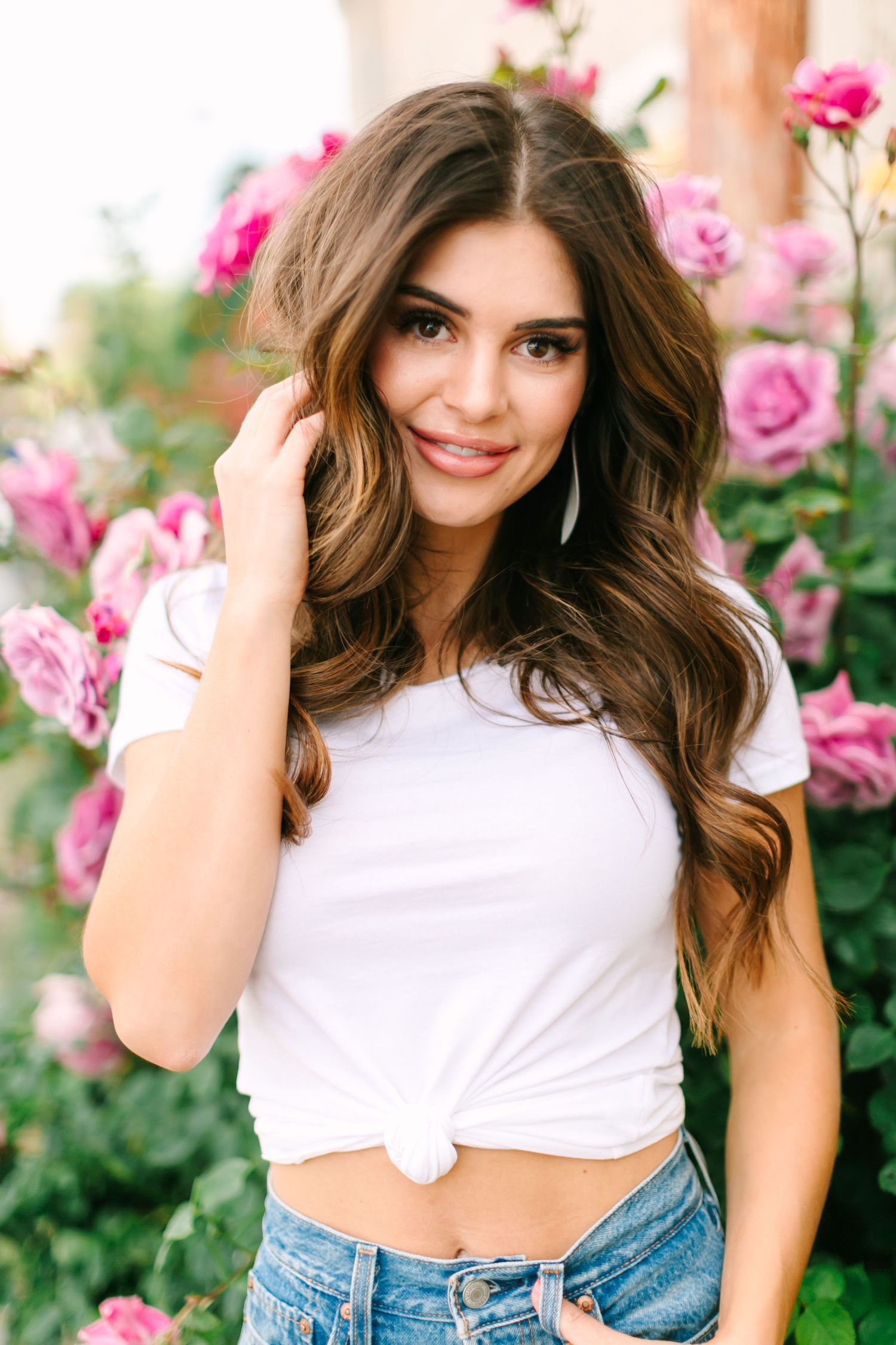 Woman in white crop top and denim poses in front of pink rose bushes in outdoor portrait session.