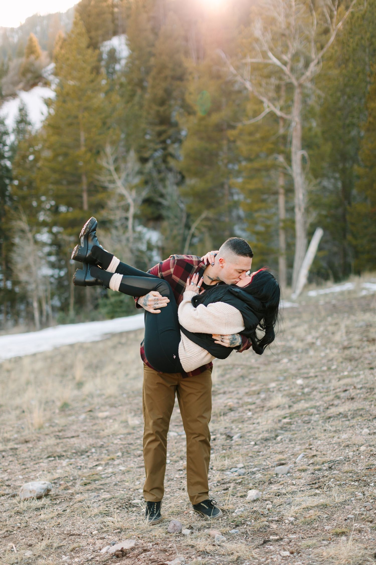 A couple embraces on a snowy mountain slope with evergreen trees and sunlight in the background.