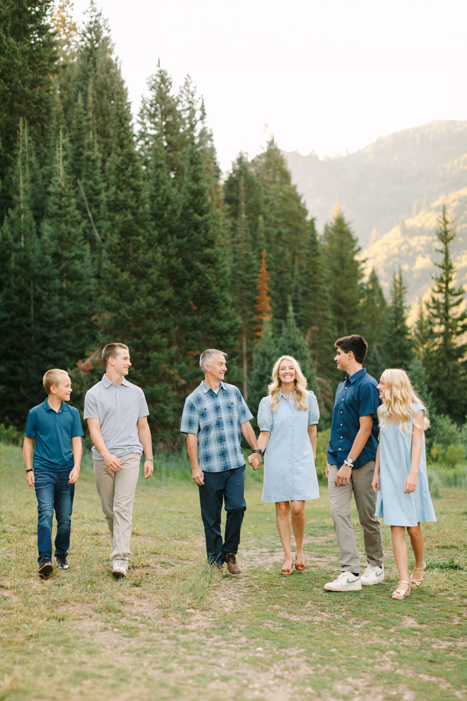 Family of six walking together in a mountain meadow surrounded by pine trees, dressed in coordinating blue and gray outfits.