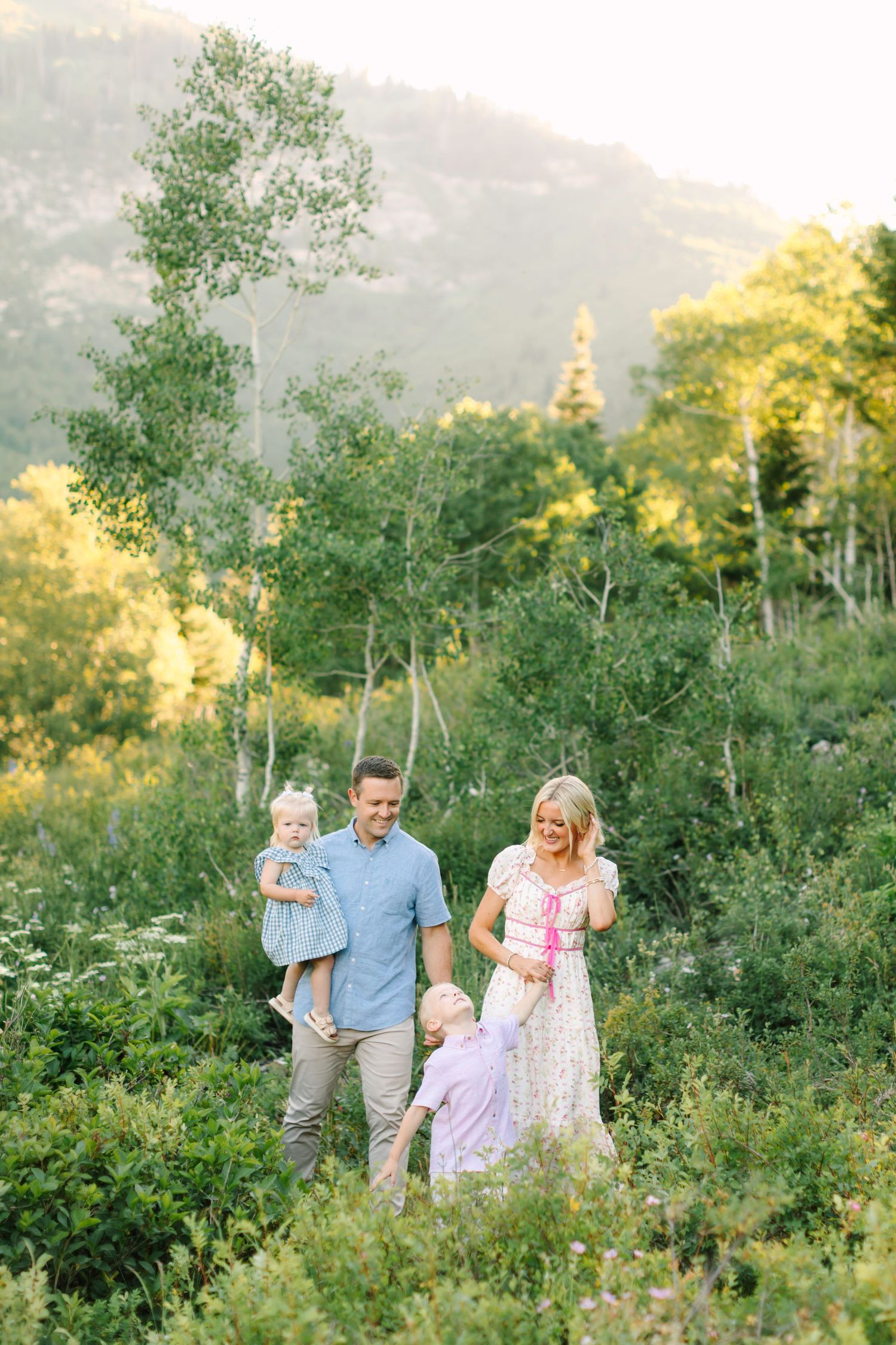 Family of four walking through lush green mountain meadow during golden hour, parents smiling with two young children.