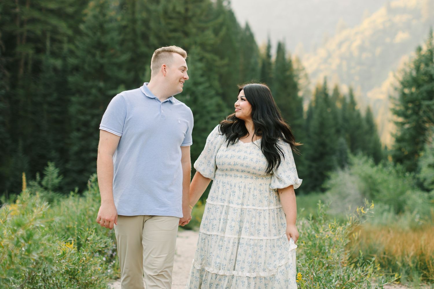 Couple holding hands smiling at each other on a mountain path surrounded by pine trees and wildflowers in Utah.