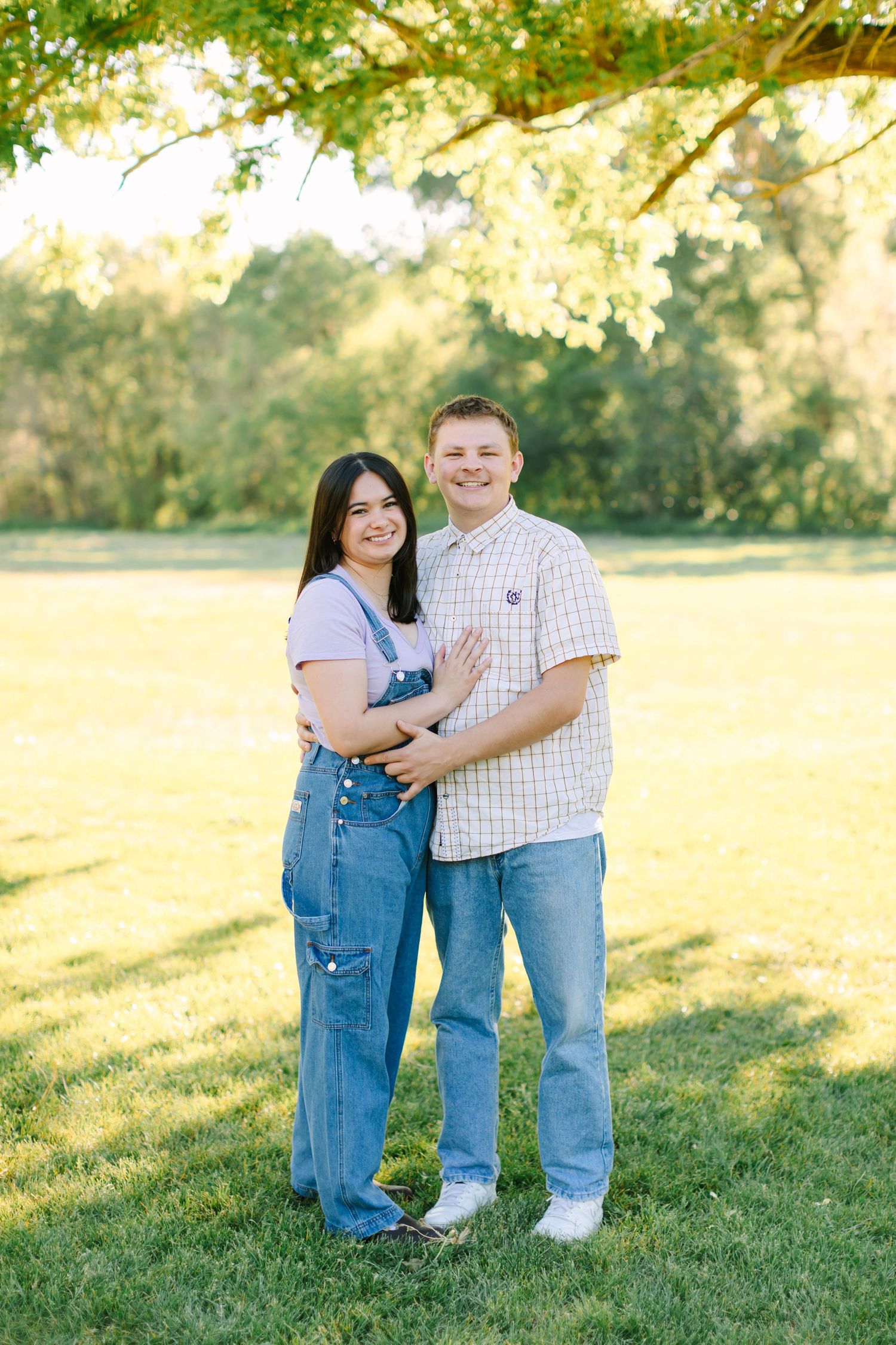 A couple in casual attire embraces while standing on green grass during golden hour sunset.
