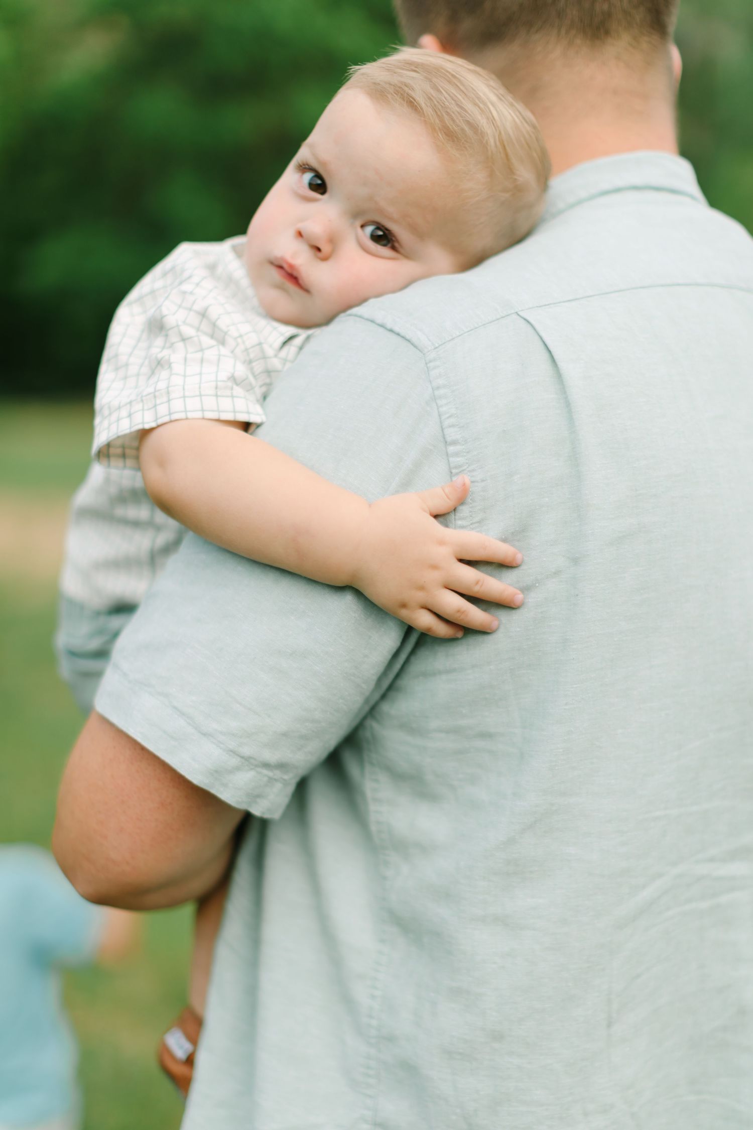 A small child in a checkered shirt being held and looking over someone's shoulder in an outdoor setting.