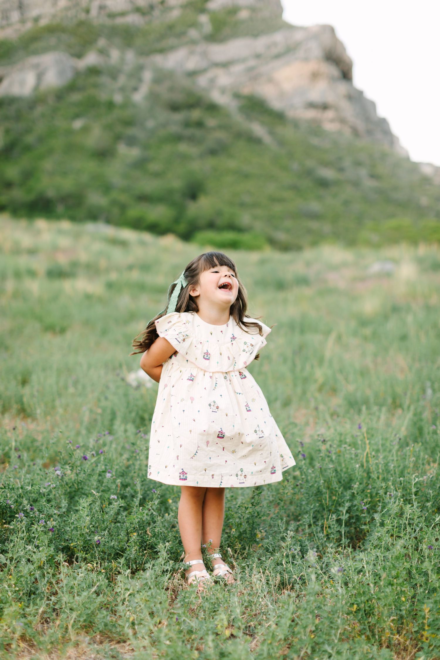 A joyful little girl in a floral dress laughs in a green meadow with a rocky mountain backdrop.