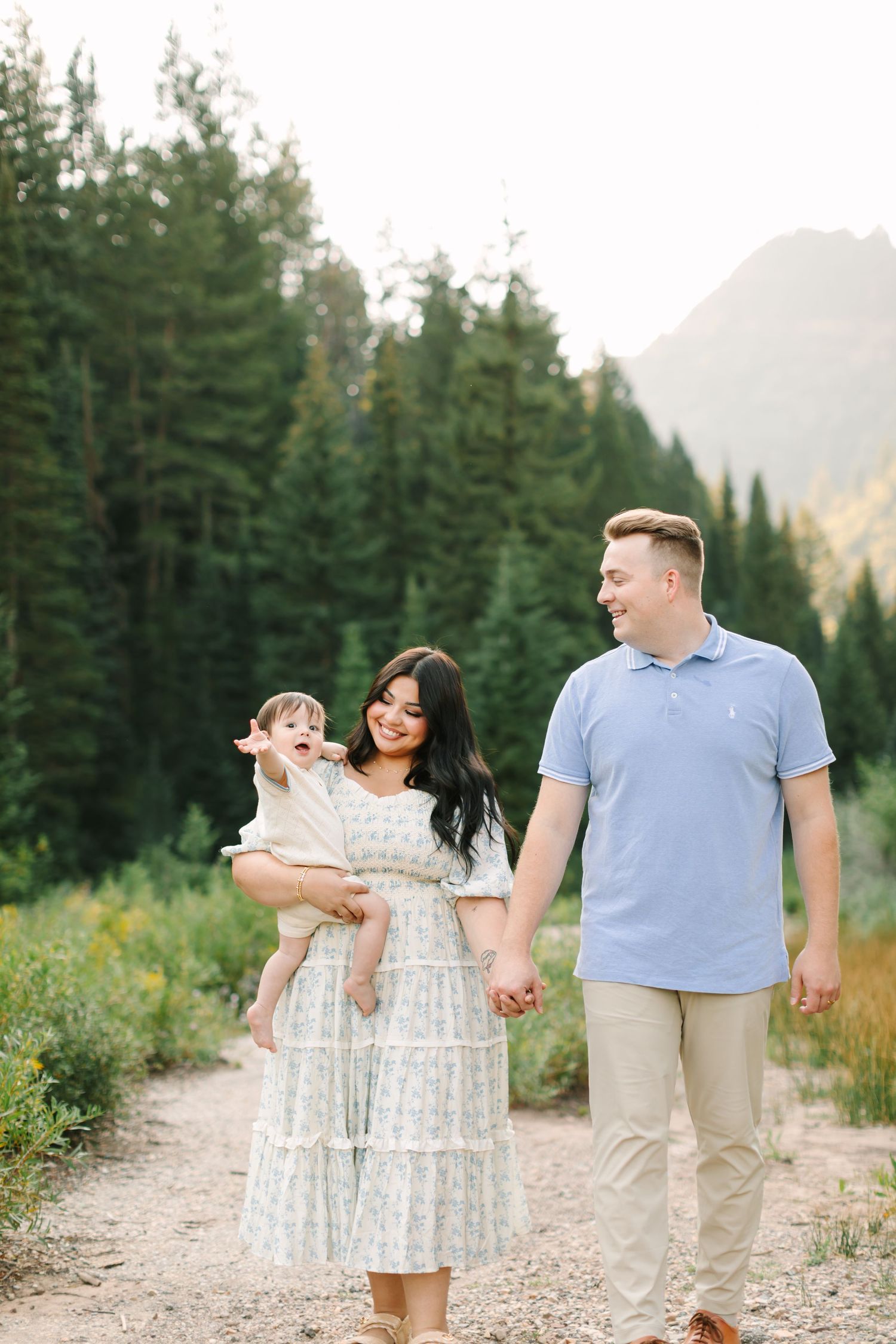 A happy family of three walks on a mountain trail, with mom holding a baby and dad smiling, surrounded by pine trees in Utah.