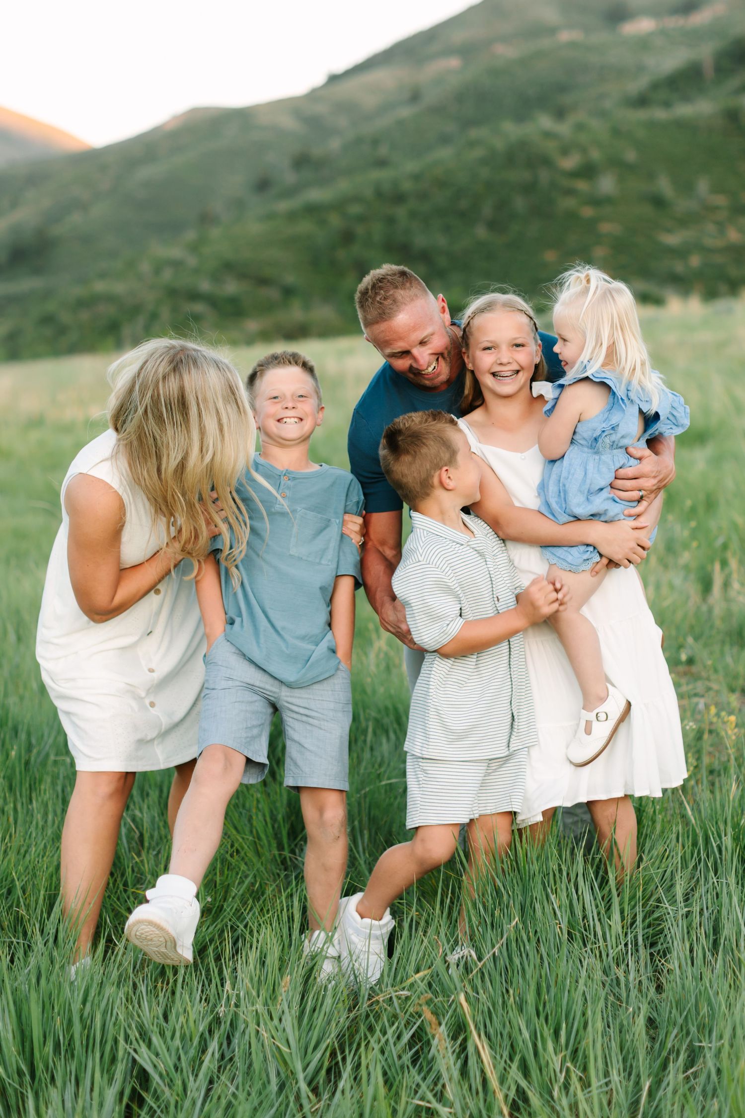 Happy family of five laughing together in a green meadow with mountain backdrop during golden hour.