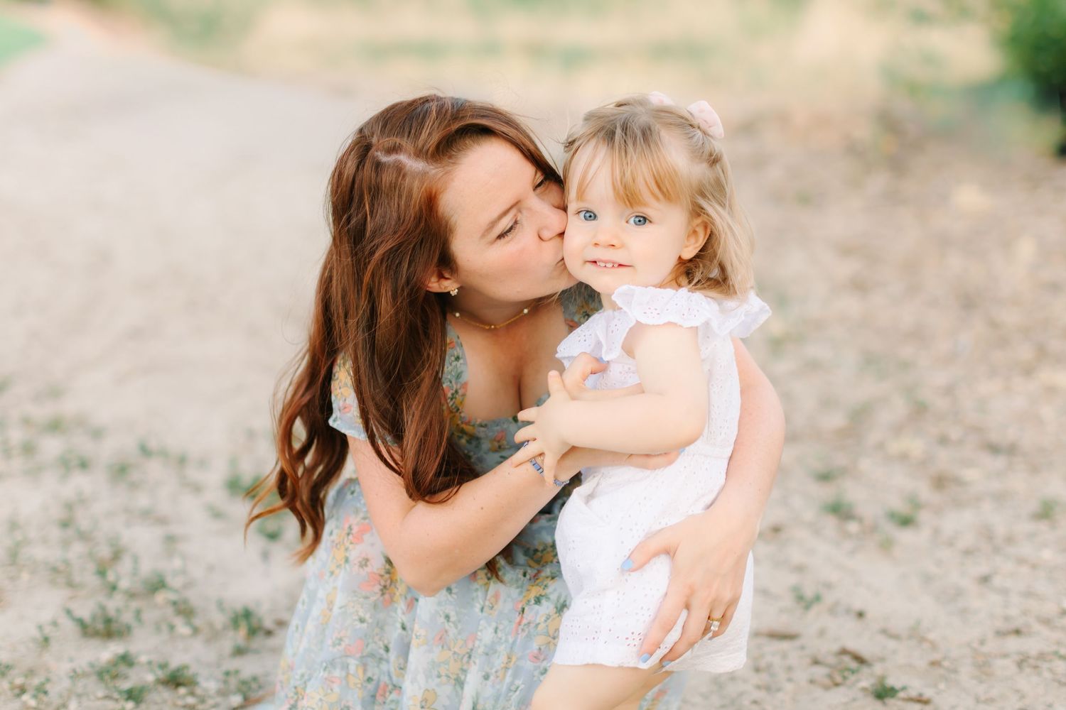 A young mother in a floral dress kisses her toddler daughter in a white dress outdoors.