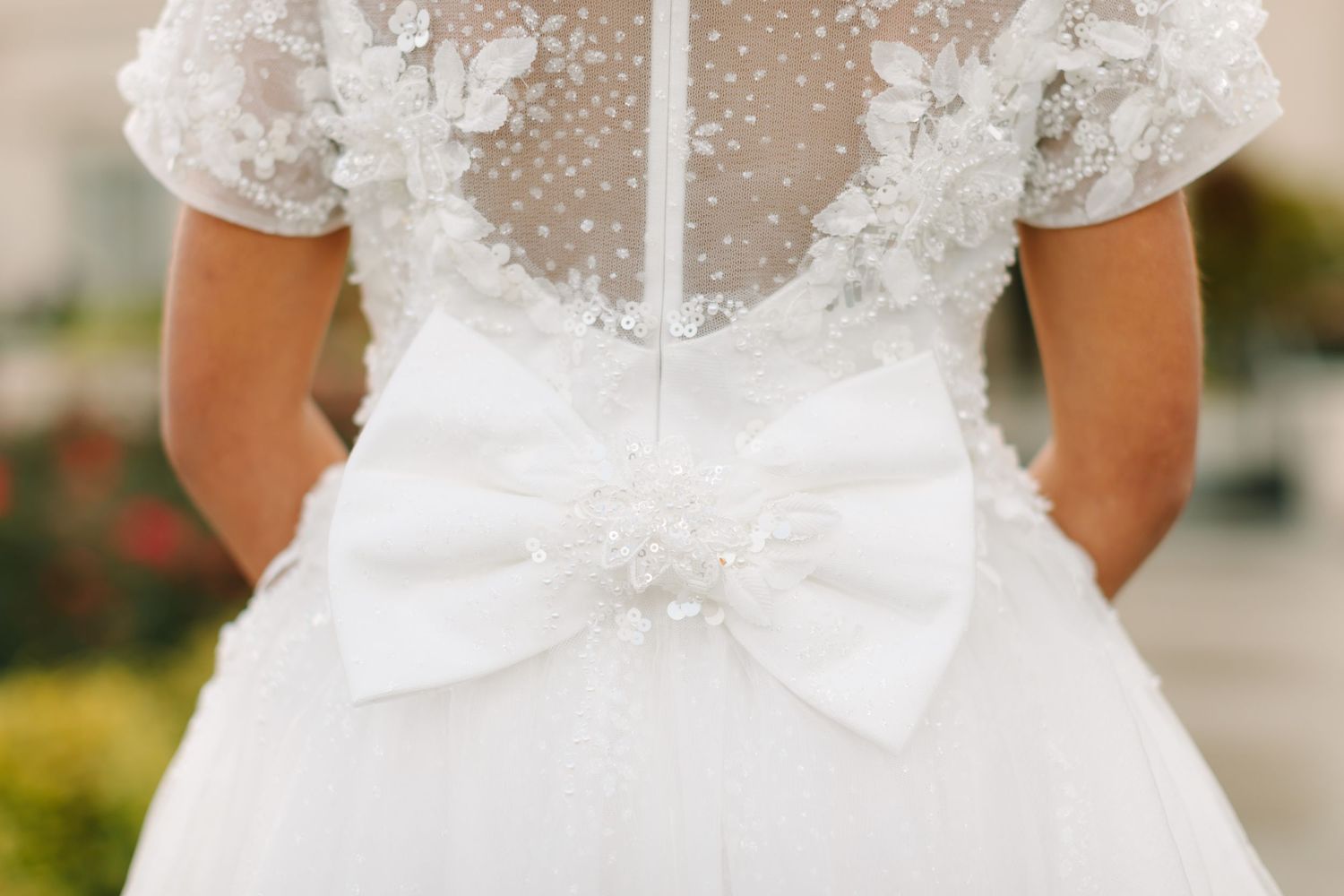 A wedding dress with intricate lace details and sheer sleeves hangs elegantly in natural light.
