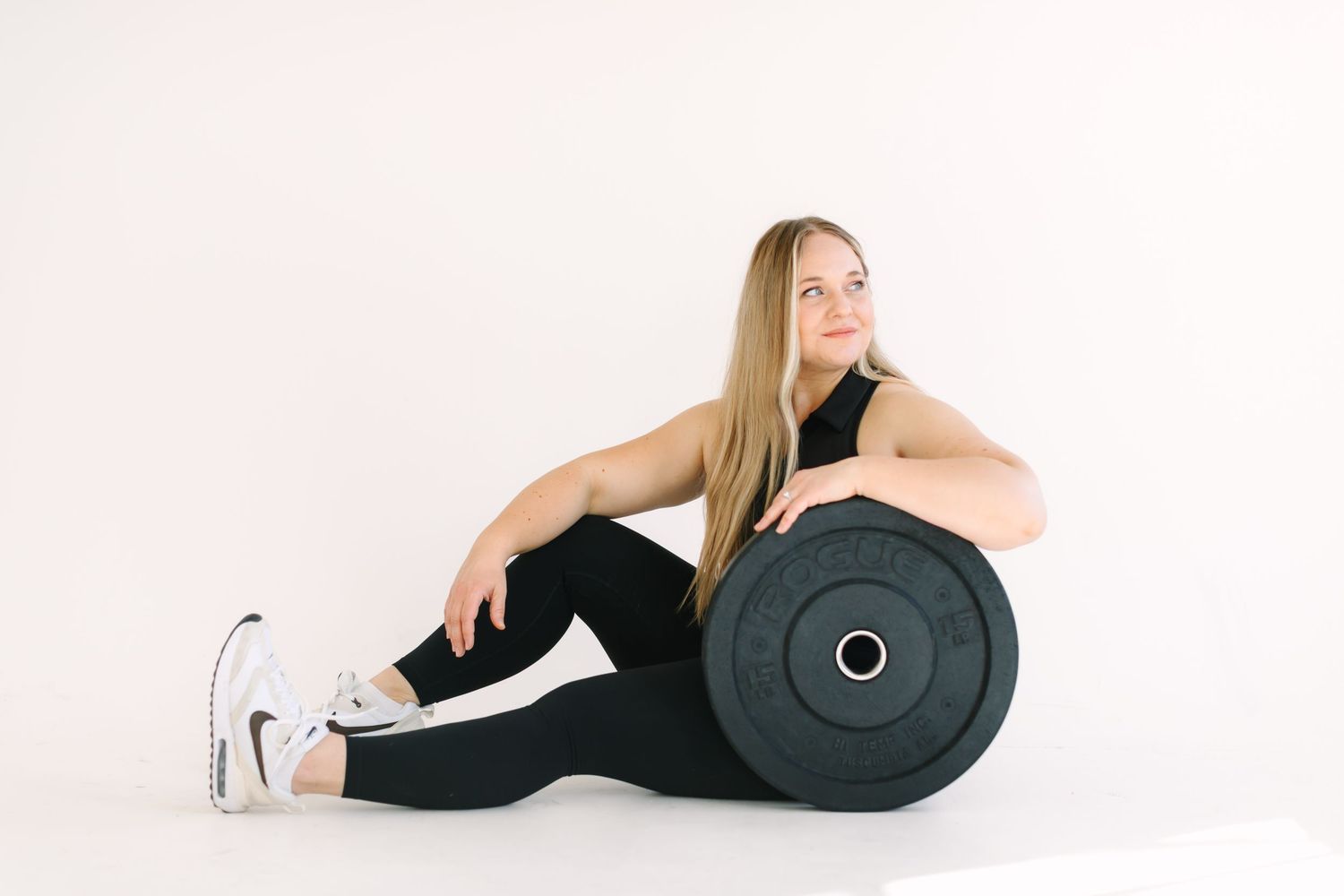 Fitness model in black athletic wear poses with large weight plate against white background.