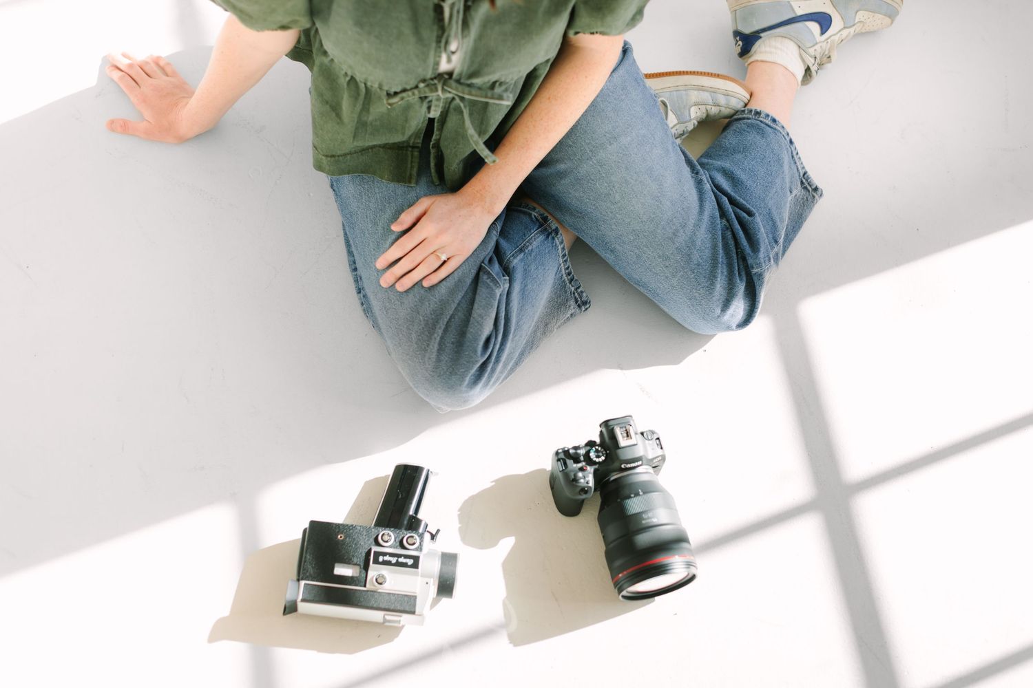 Person wearing olive green shirt and blue jeans sitting cross-legged on white floor in natural lighting.