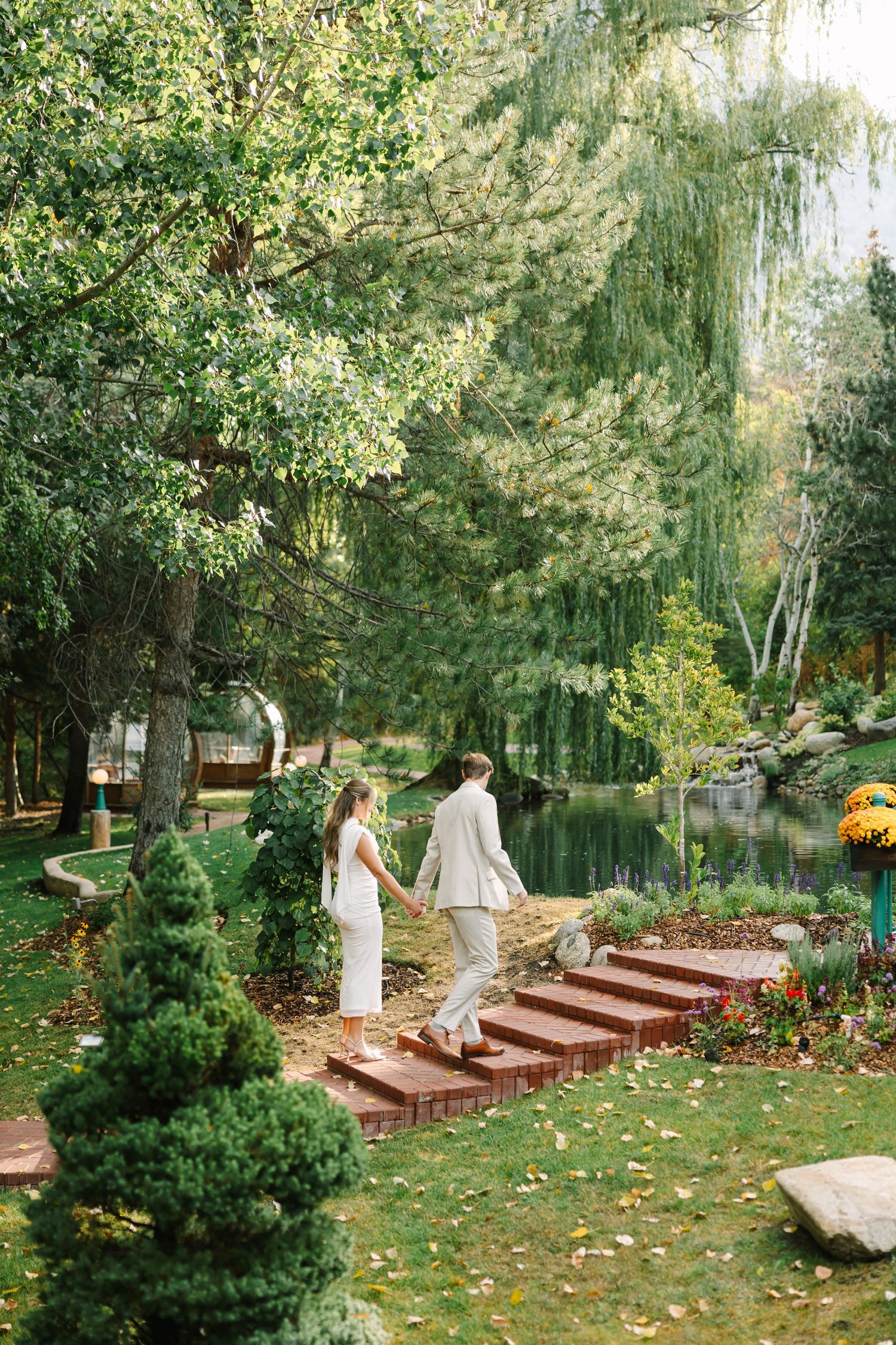 Two people descend wooden steps in a serene garden setting with weeping willows and a pond in the background.