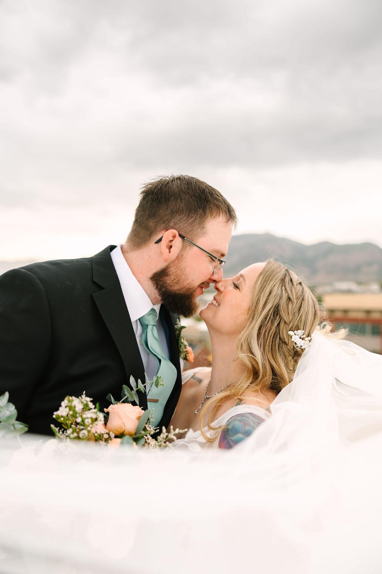 A romantic wedding couple shares a tender kiss against a cloudy mountain backdrop with white veil flowing in the breeze.