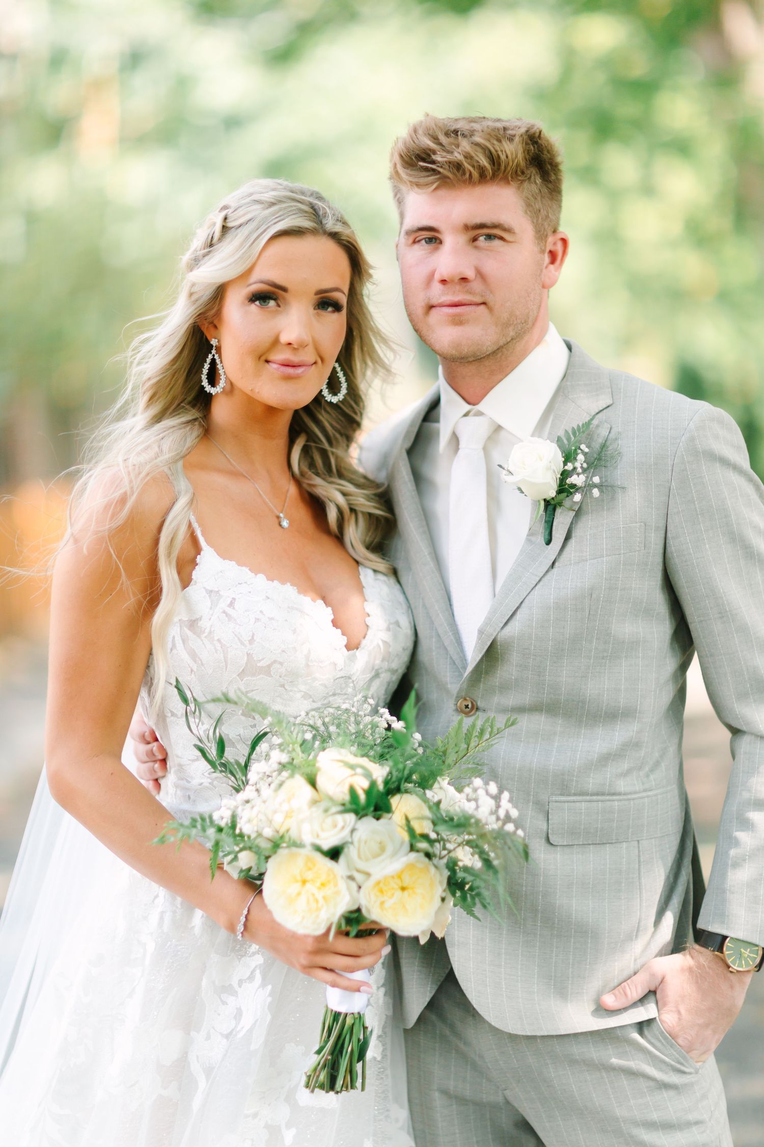 A wedding couple poses outdoors in formal attire with white floral bouquet against a soft green natural background.