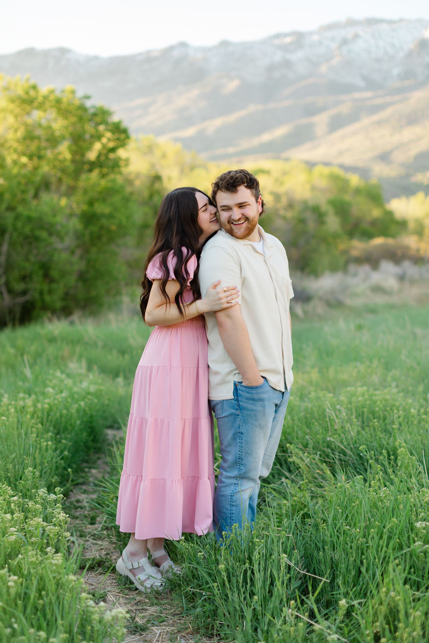 Playful couple sharing tender moments in a spring meadow with mountains visible behind them for their engagement photoshoot in Alpine, Utah.