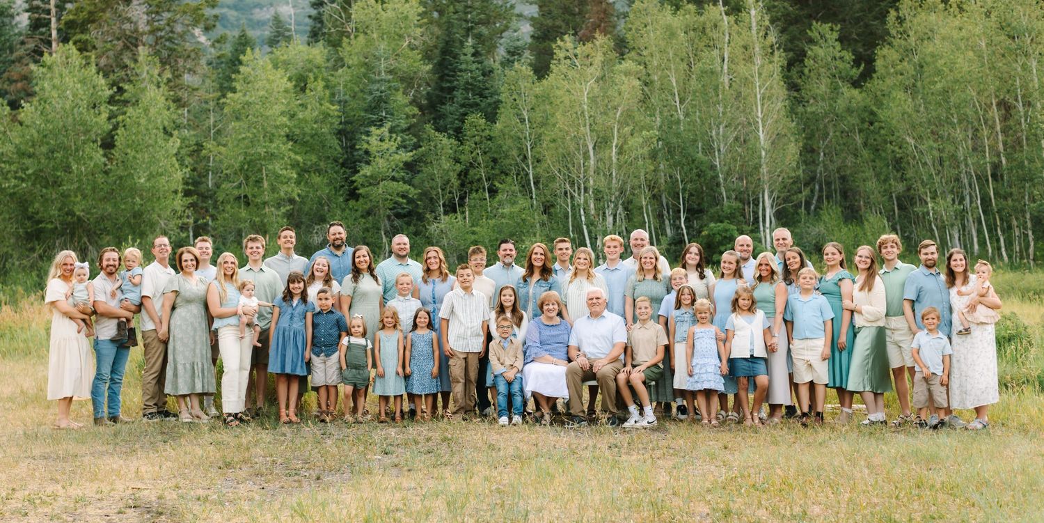 Large multigenerational family portrait outdoors in a mountain meadow with lush green aspen trees in the background in Provo, Utah.