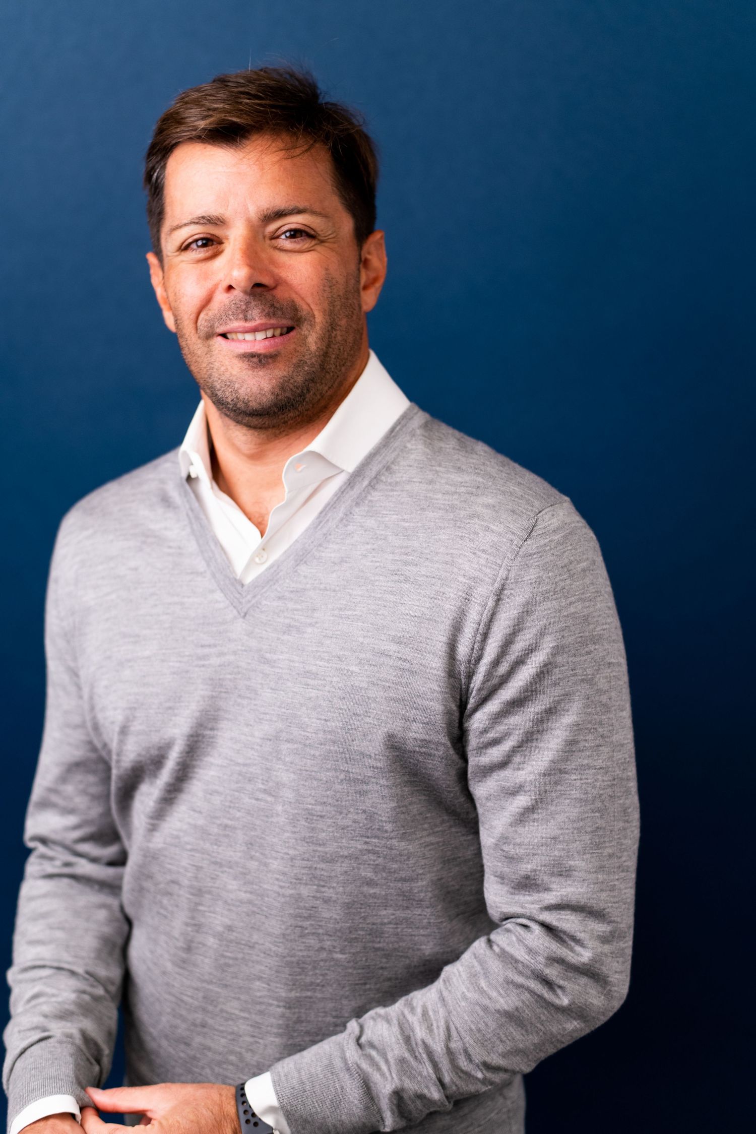 Male business professional wearing a gray sweater over a collared shirt, photographed against a blue backdrop for a professional headshot.