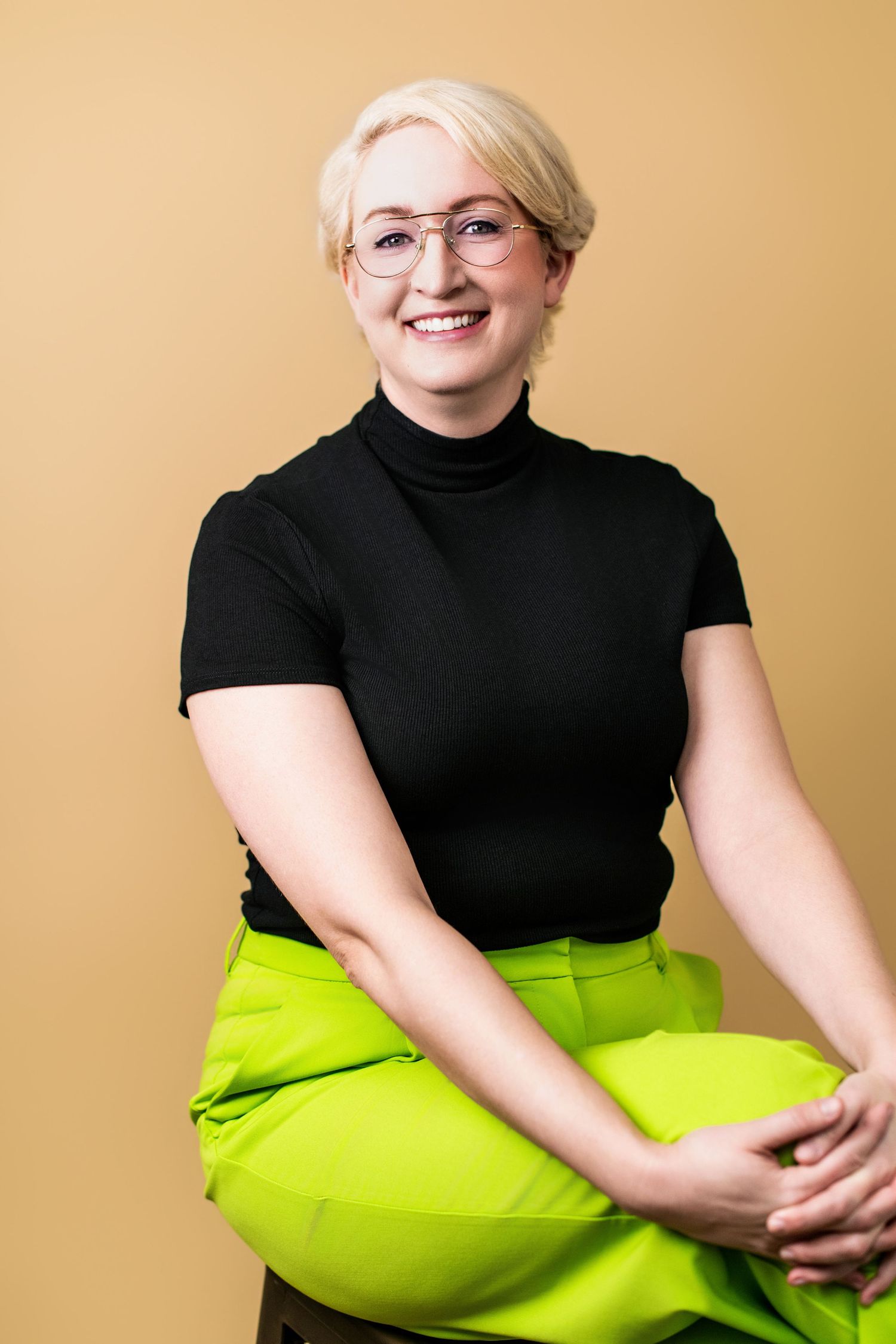 Modern business headshot of a woman with short blonde hair and glasses, wearing a black top against a warm yellow studio backdrop.