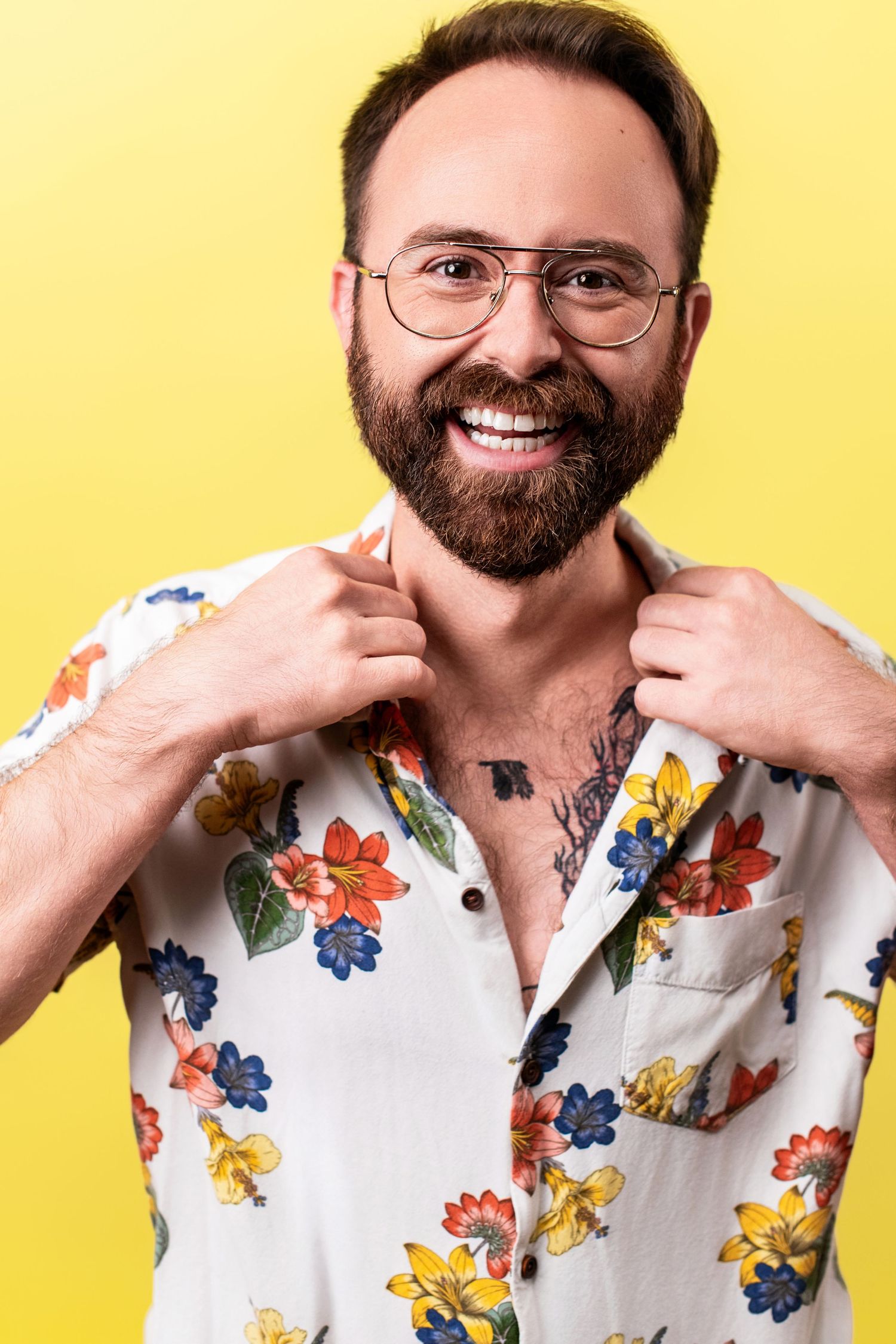Smiling bearded man in glasses wearing a floral shirt against a yellow background, photographed by Nice Headshots.