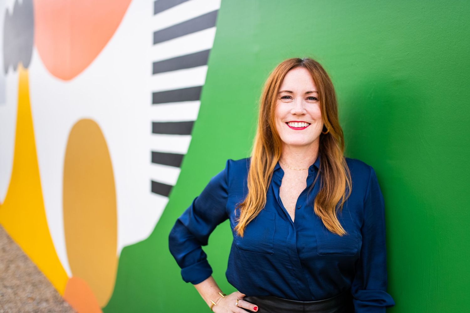 Professional female business headshot of a red-haired woman in a navy blouse, smiling confidently against a colorful mural backdrop.