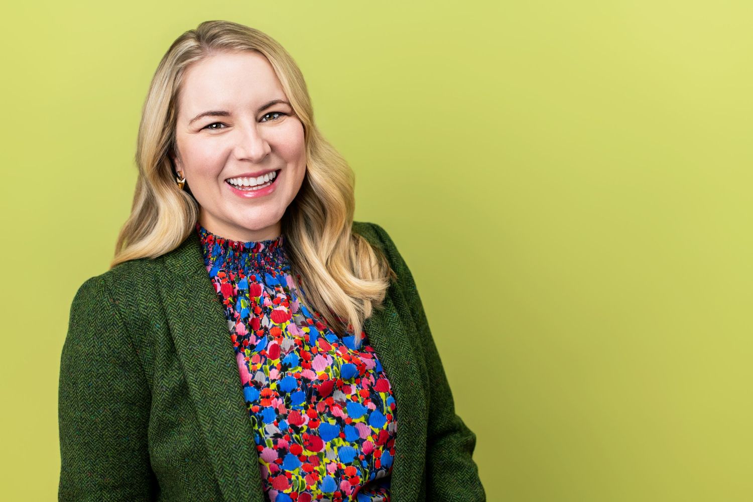 Professional business headshot of a woman in a green blazer and patterned blouse, smiling confidently against a soft green studio backdrop.
