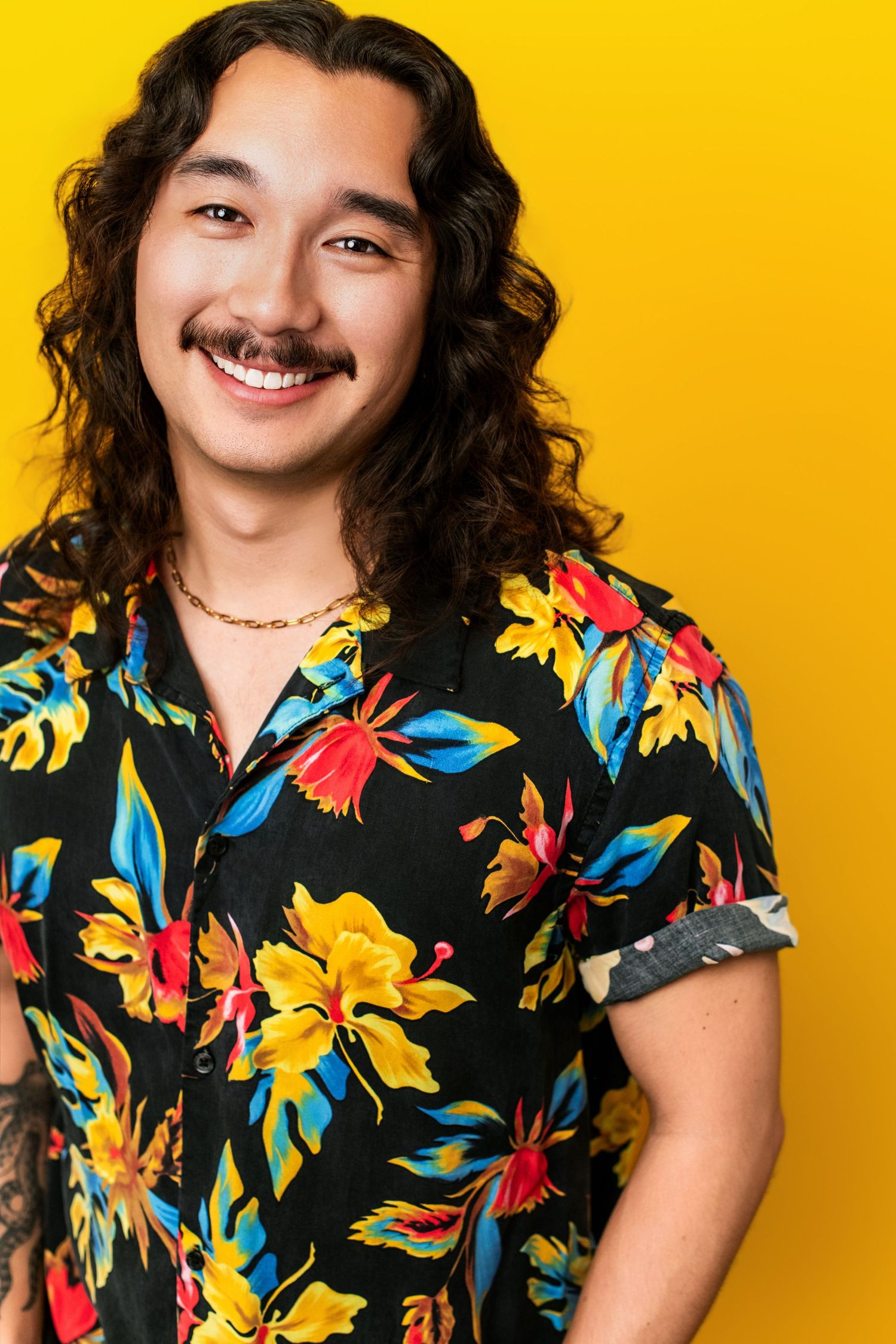 Commercial actor headshot of a smiling young adult with long hair and mustache on a bold yellow backdrop in Los Angeles.