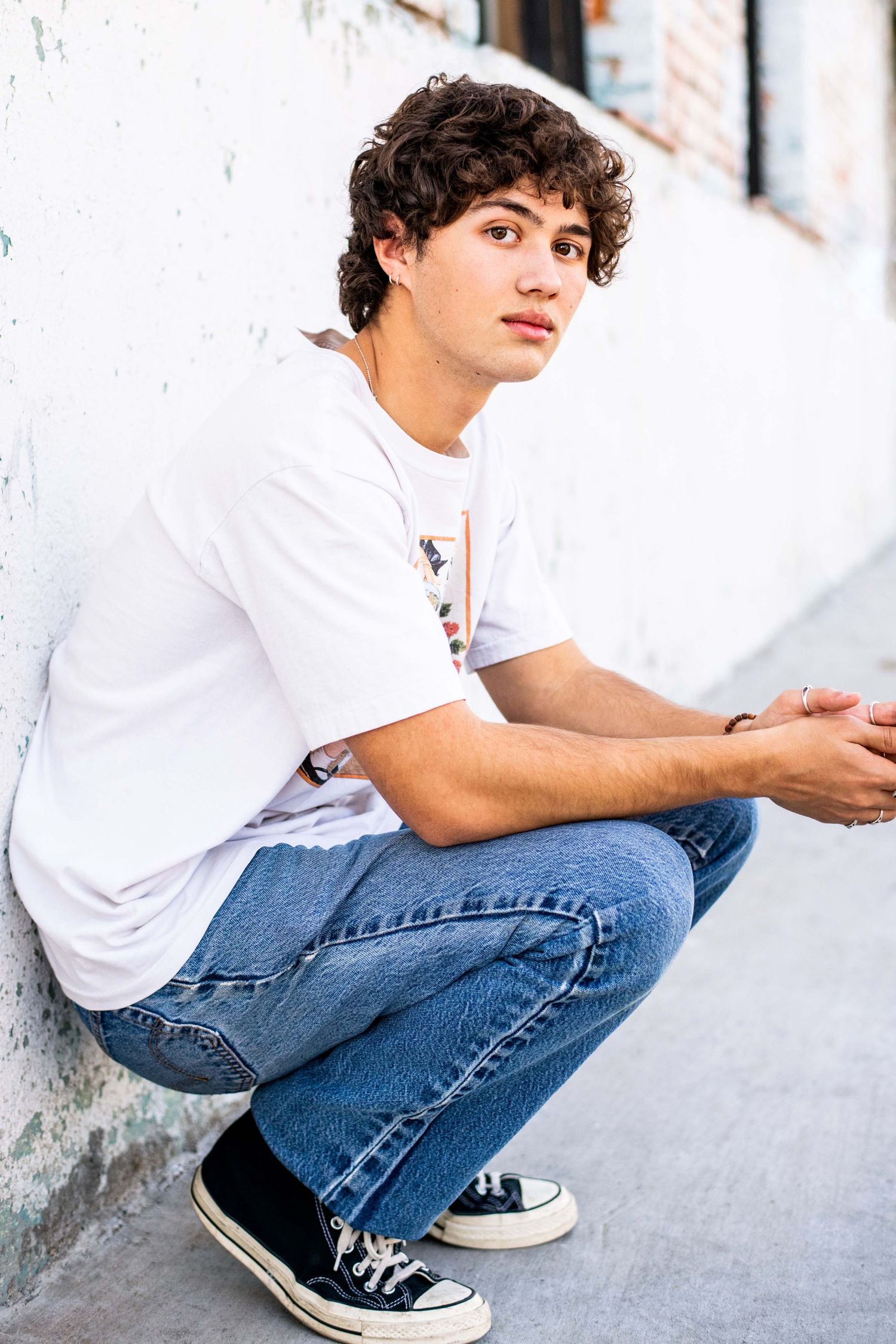 Male model crouching against a white wall wearing a casual t-shirt and jeans, photographed for a lifestyle modeling headshot in Los Angeles.